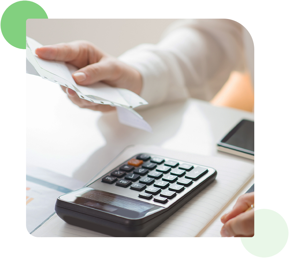 A person holding a receipt over a calculator on a desk, with a smartphone and notepad nearby.