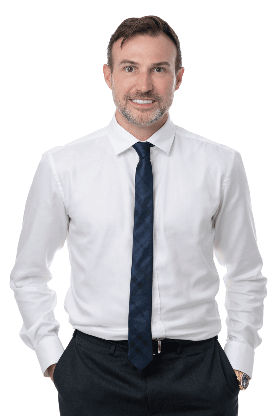 A man with light skin, brown hair, and a beard wearing a white dress shirt, navy blue tie, and black pants, standing confidently with his hands in his pockets.