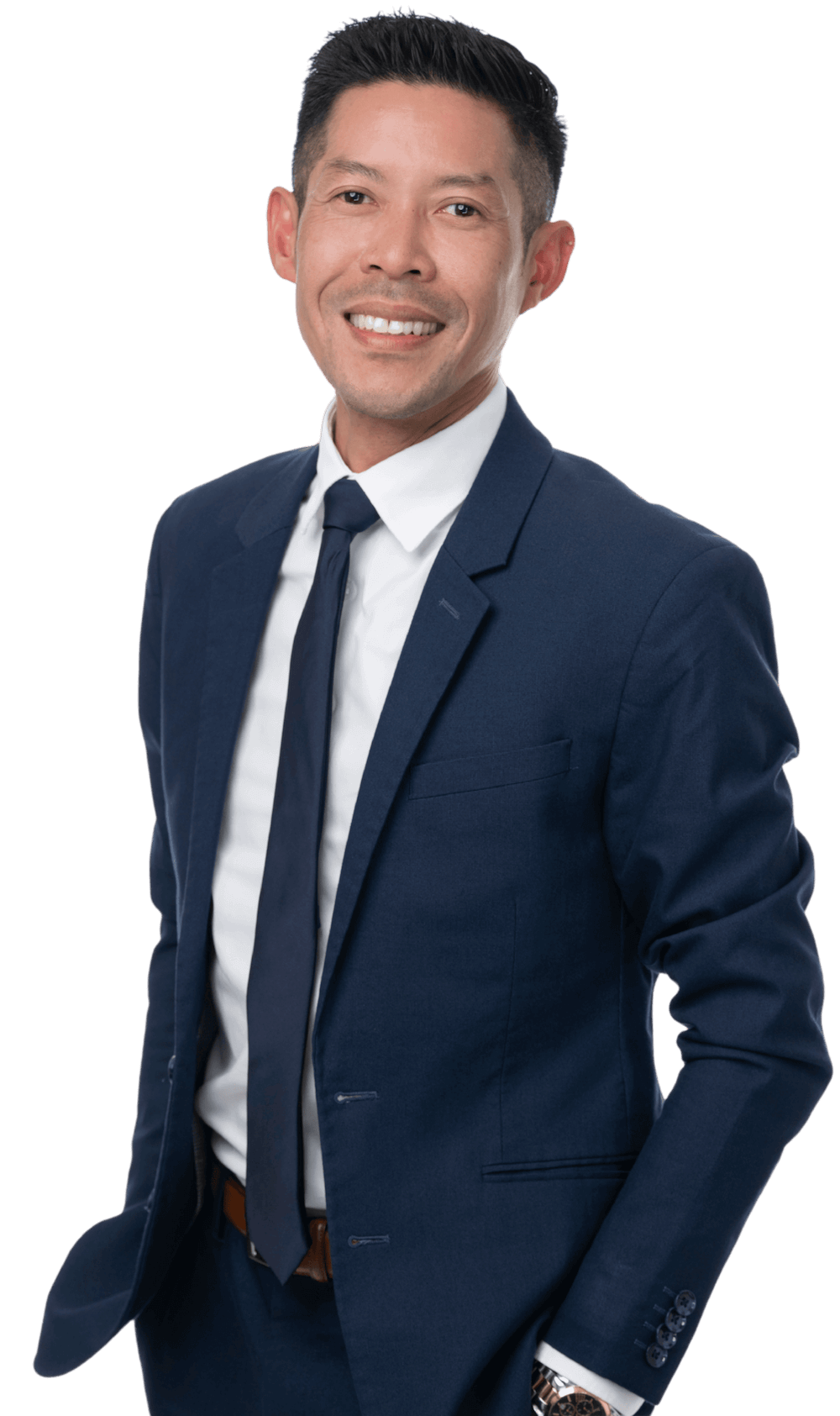 A smiling man in a navy blue suit, white shirt, and navy tie, standing with one hand in his pocket against a black background.