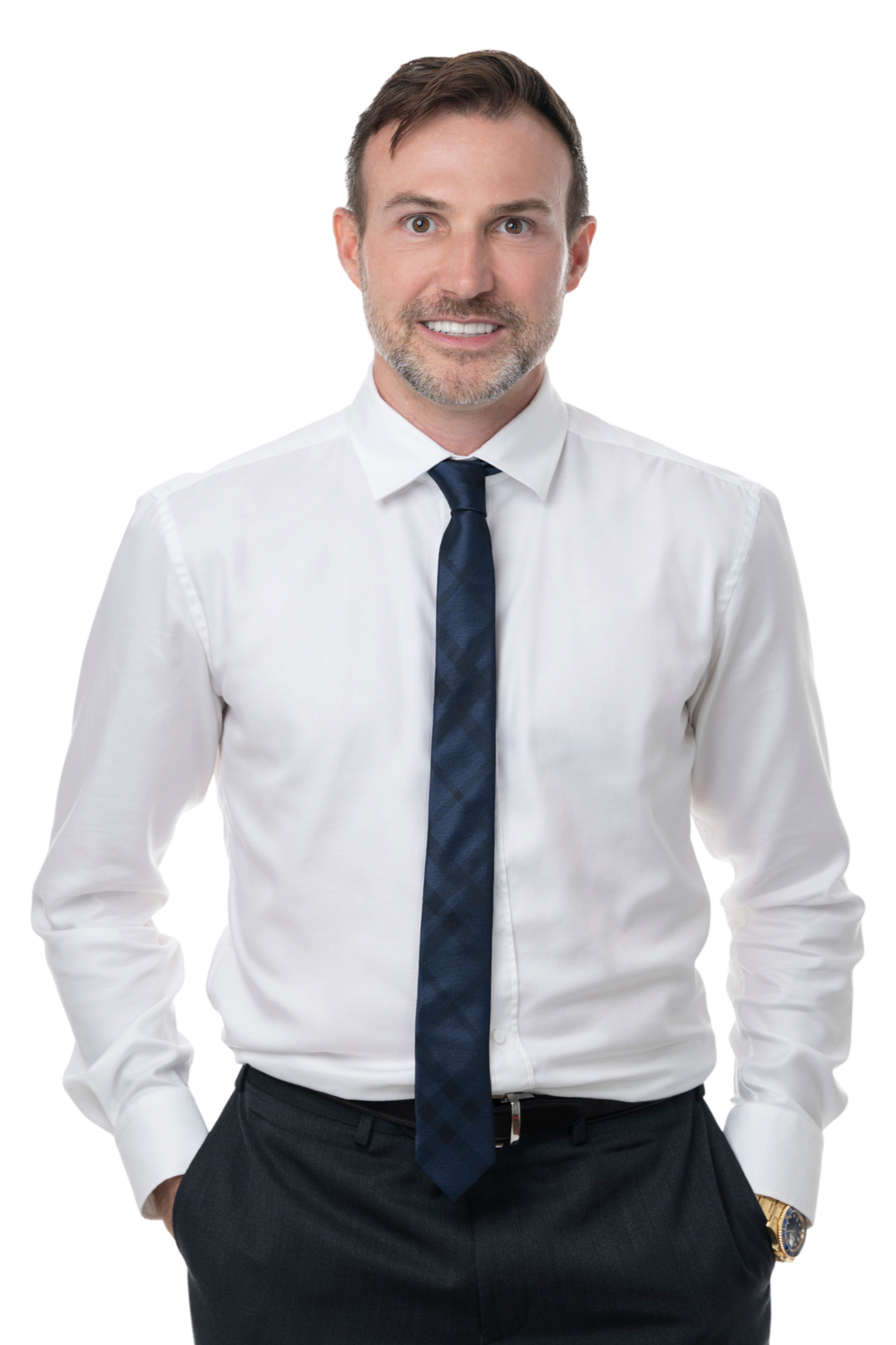 A man in a white dress shirt, navy tie, and black pants, smiling and standing with his hands in his pockets.