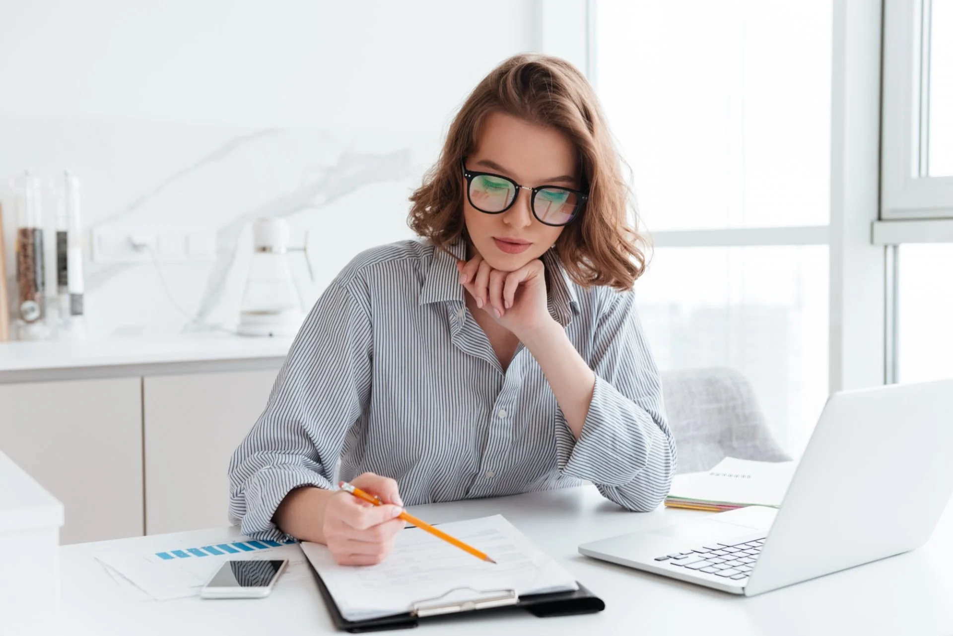 Young woman with glasses working at desk with documents, laptop, and smartphone in bright office.