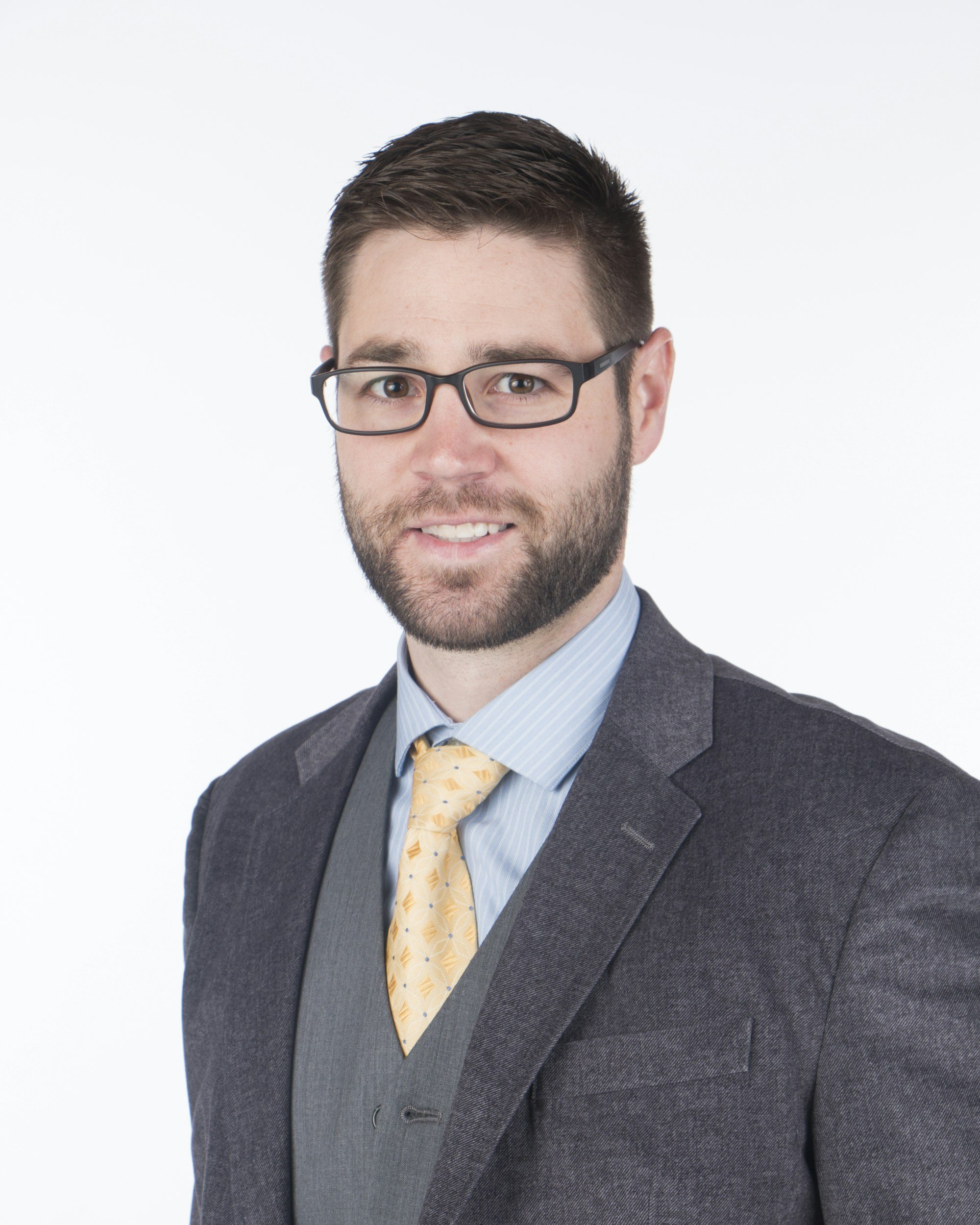 A man with a beard and short brown hair, wearing a navy suit, white shirt, and navy tie, smiling against a light gray background.