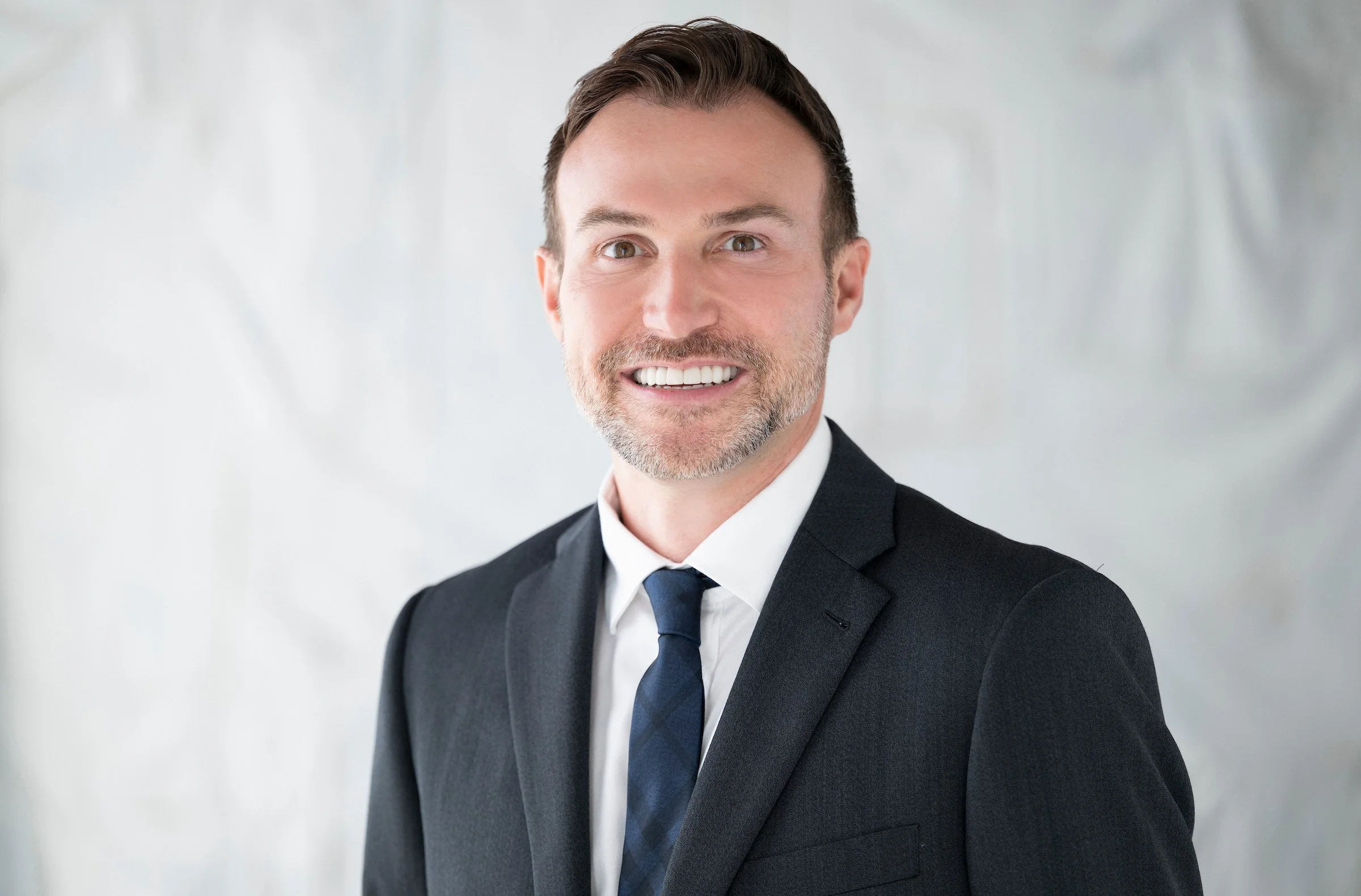 A man in a dark suit, white shirt, and patterned tie sitting on a black stool against a gray background, smiling and wearing a watch on his left wrist.