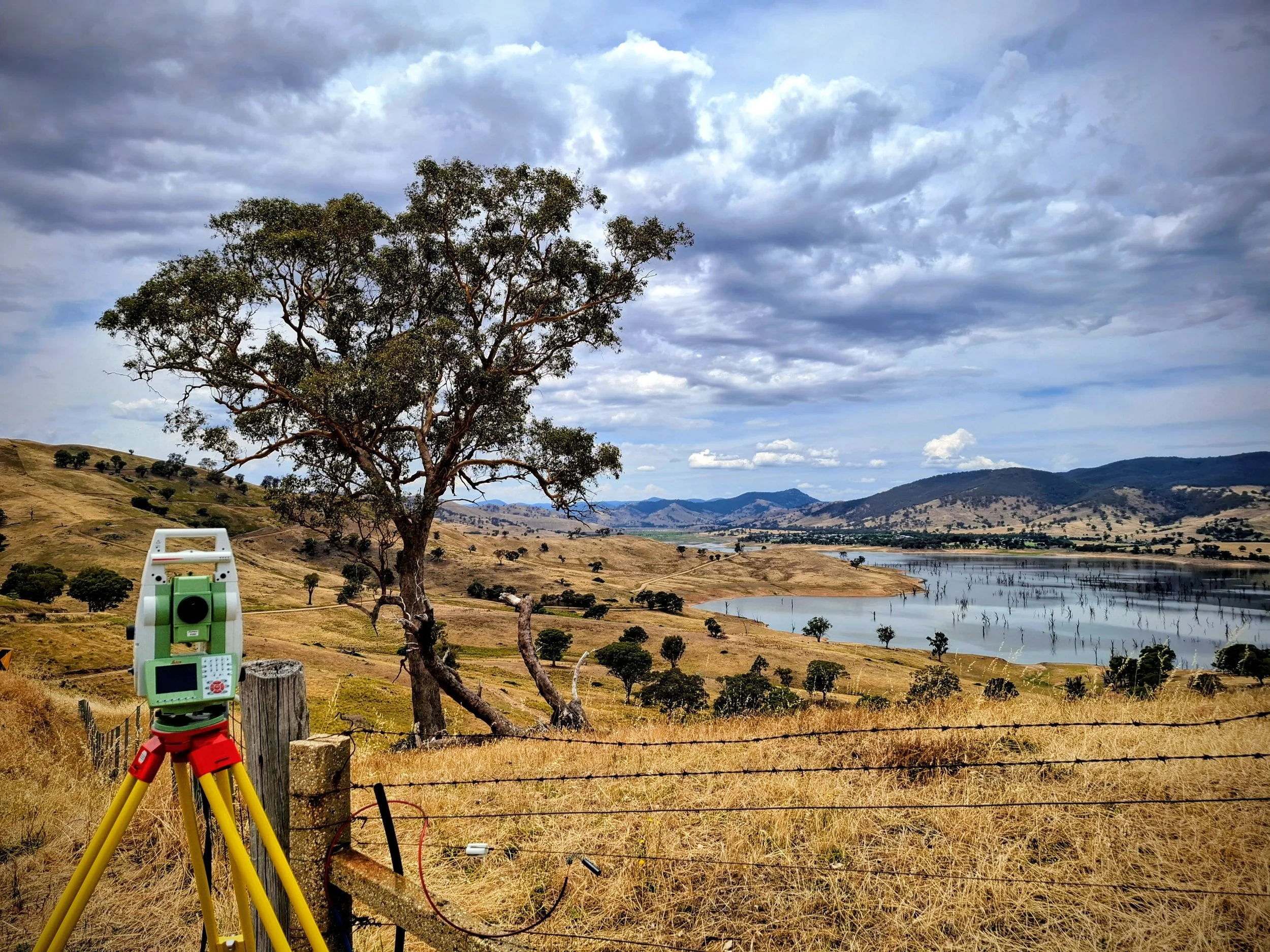 Surveying instrument set up on a yellow tripod in a dry, grassy field with a barbed wire fence, overlooking a large tree, hills, and Lake Hume under a cloudy sky.