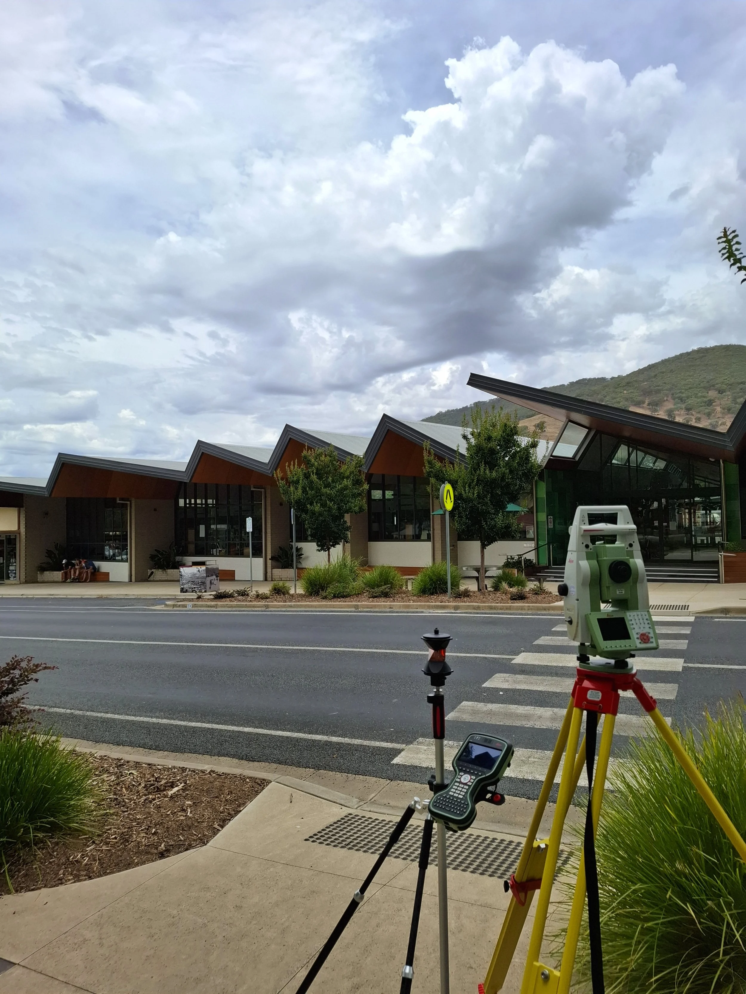 Surveying equipment set up on a sidewalk, with a building featuring a distinctive roof design, trees, and hills in the background. 