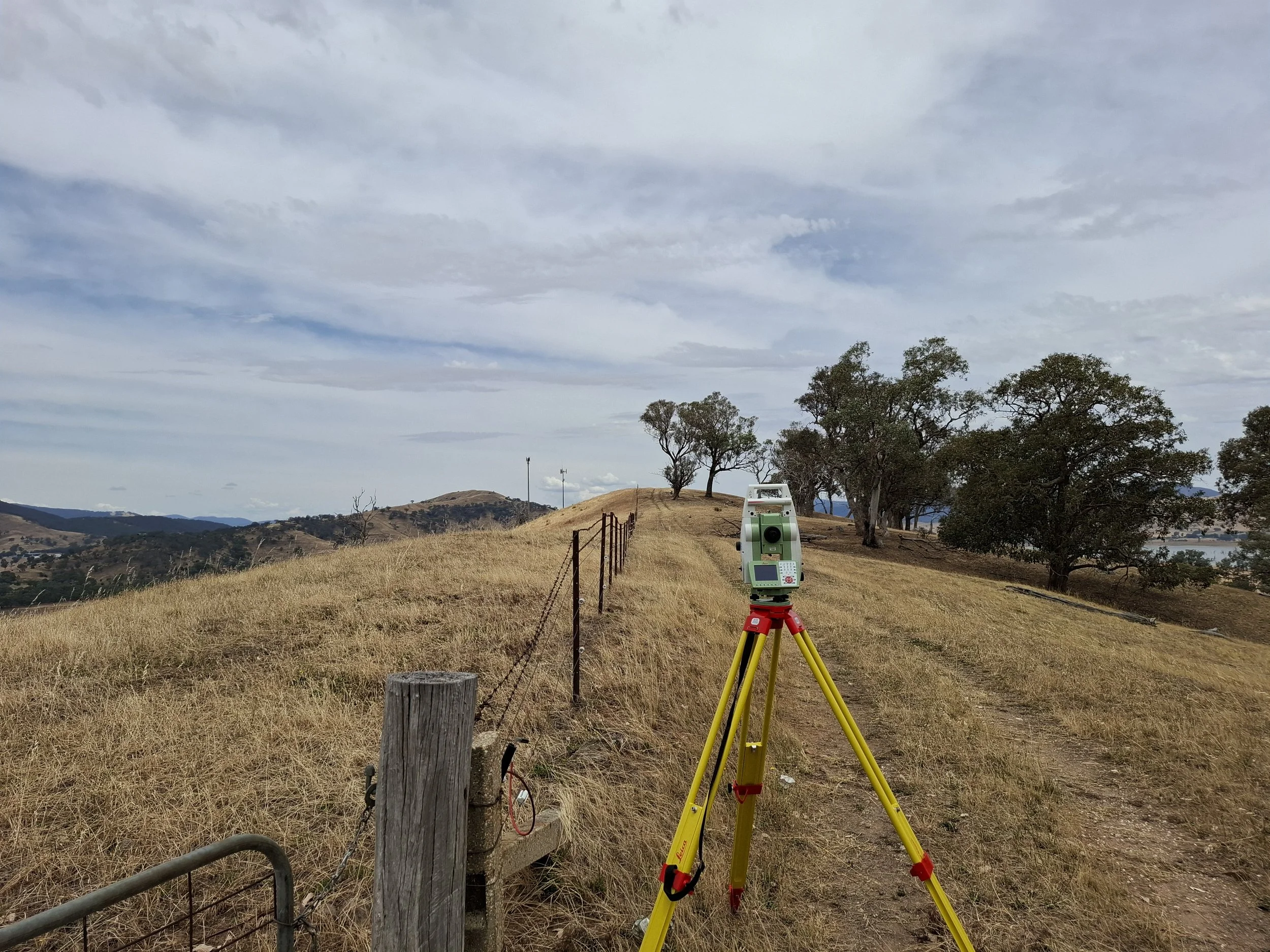 A landscape with rolling hills, trees, and a cloudy sky. There is a surveying station with a green and white instrument on a yellow tripod in the foreground, and a fence along a grassy path. In the background telecommunications towers are visible.