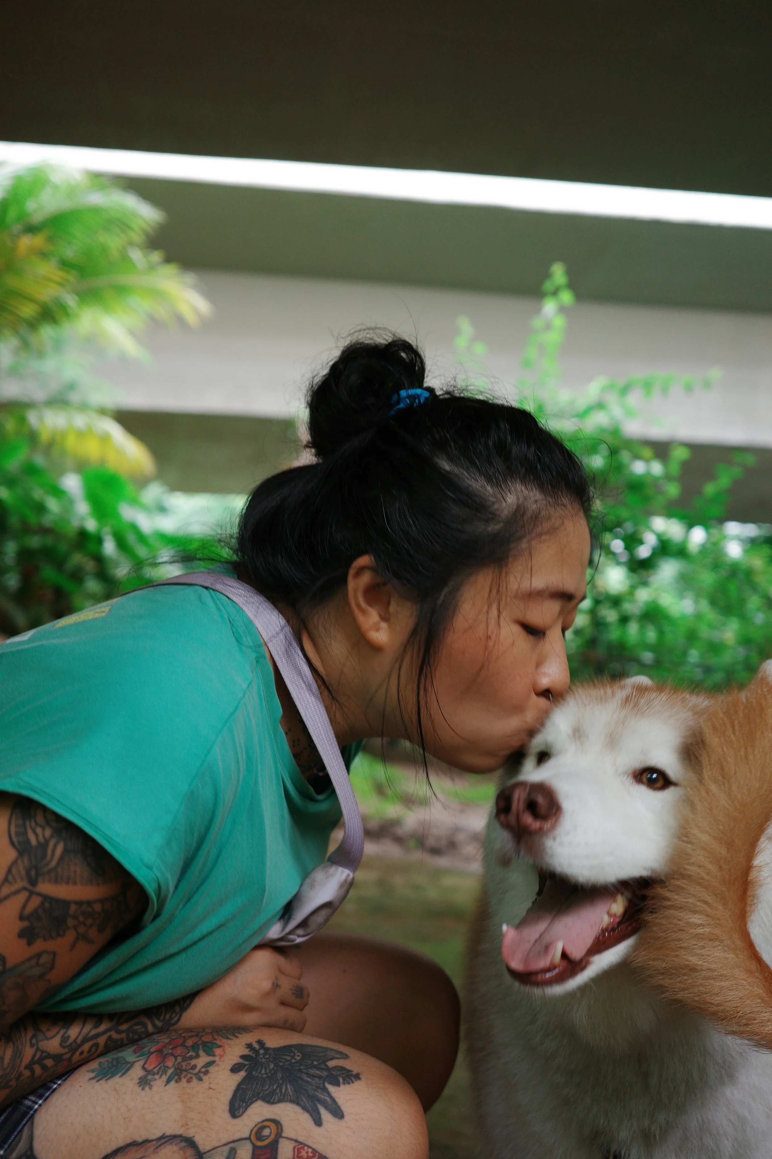 A woman with tattoos kissing a husky dog on the nose in a lush outdoor setting.