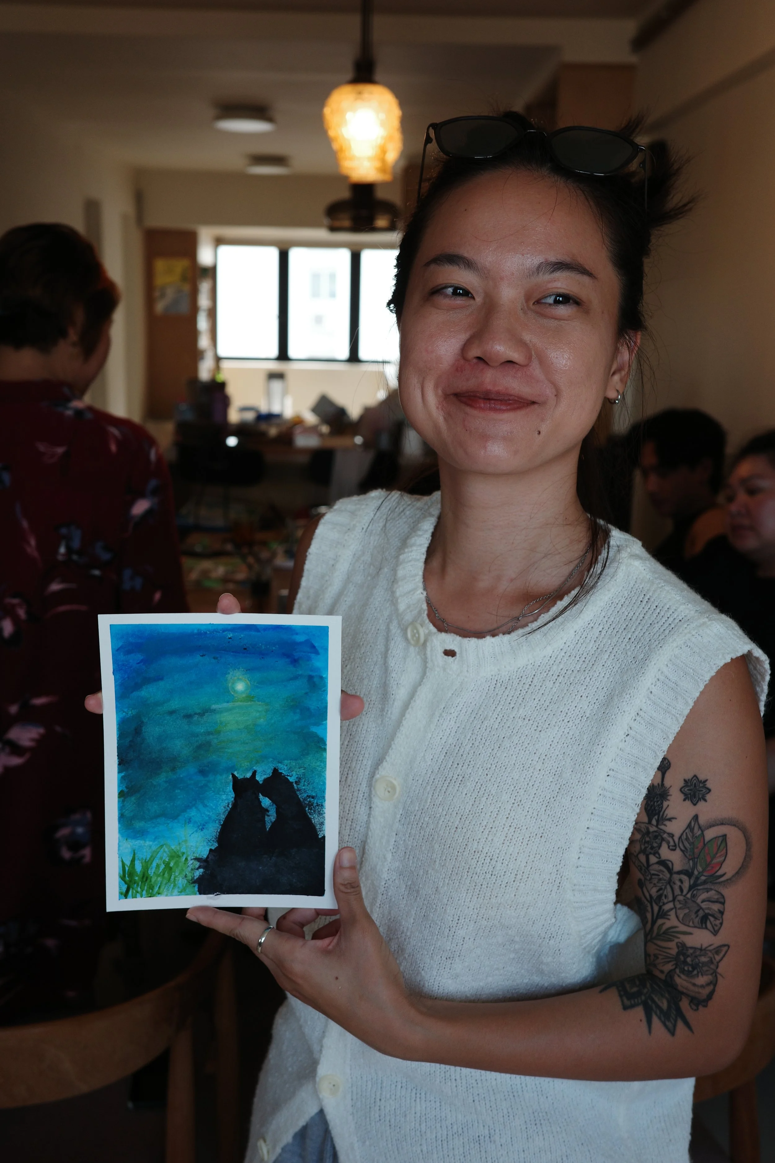 A woman with tattoos on her arm and glasses on her head holds a watercolor painting of two cats sitting near water under a moonlit sky at what appears to be an indoor art gathering.