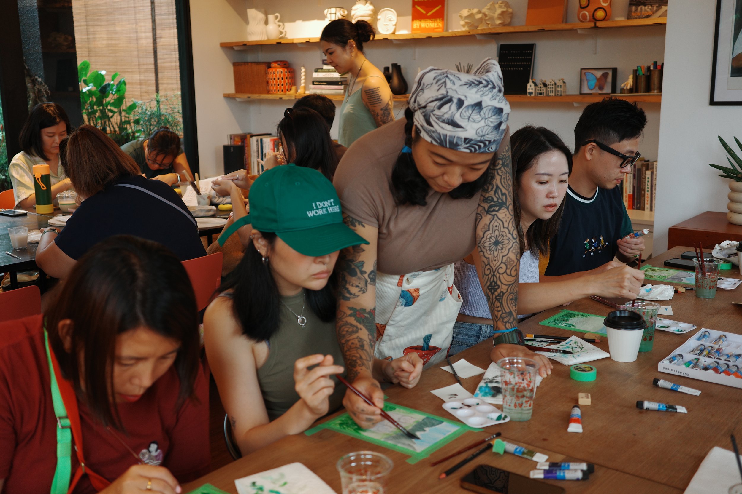 People gathered at a table engaged in watercolor painting activity, with art supplies, paint tubes, brushes, and water glasses on the table, and shelves with books and decorative items in the background.