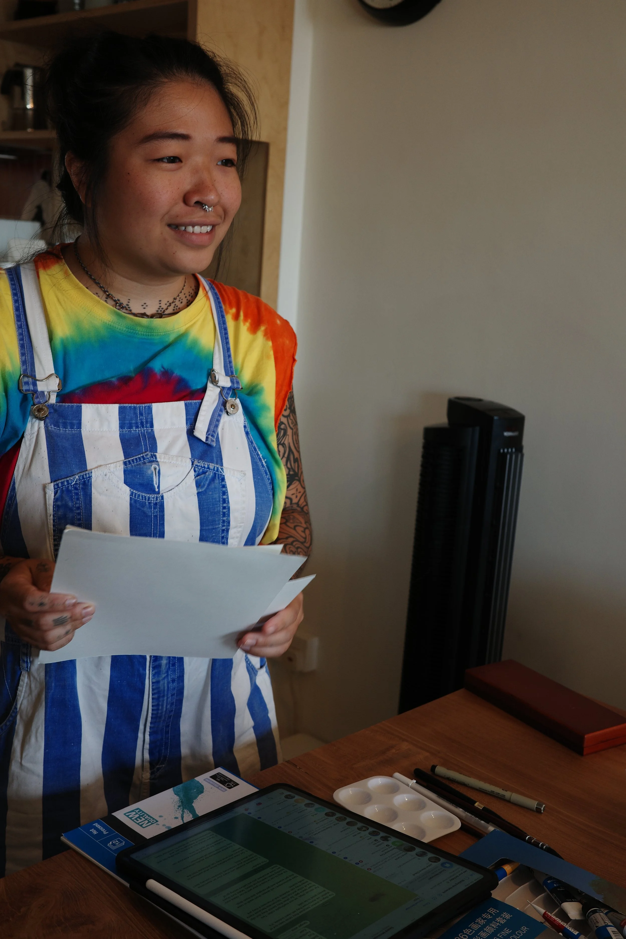 A woman with dark hair wearing a rainbow tie-dye shirt and a blue and white striped apron, holding a piece of paper, standing at a table with an iPad, paint palette, brushes, and markers.