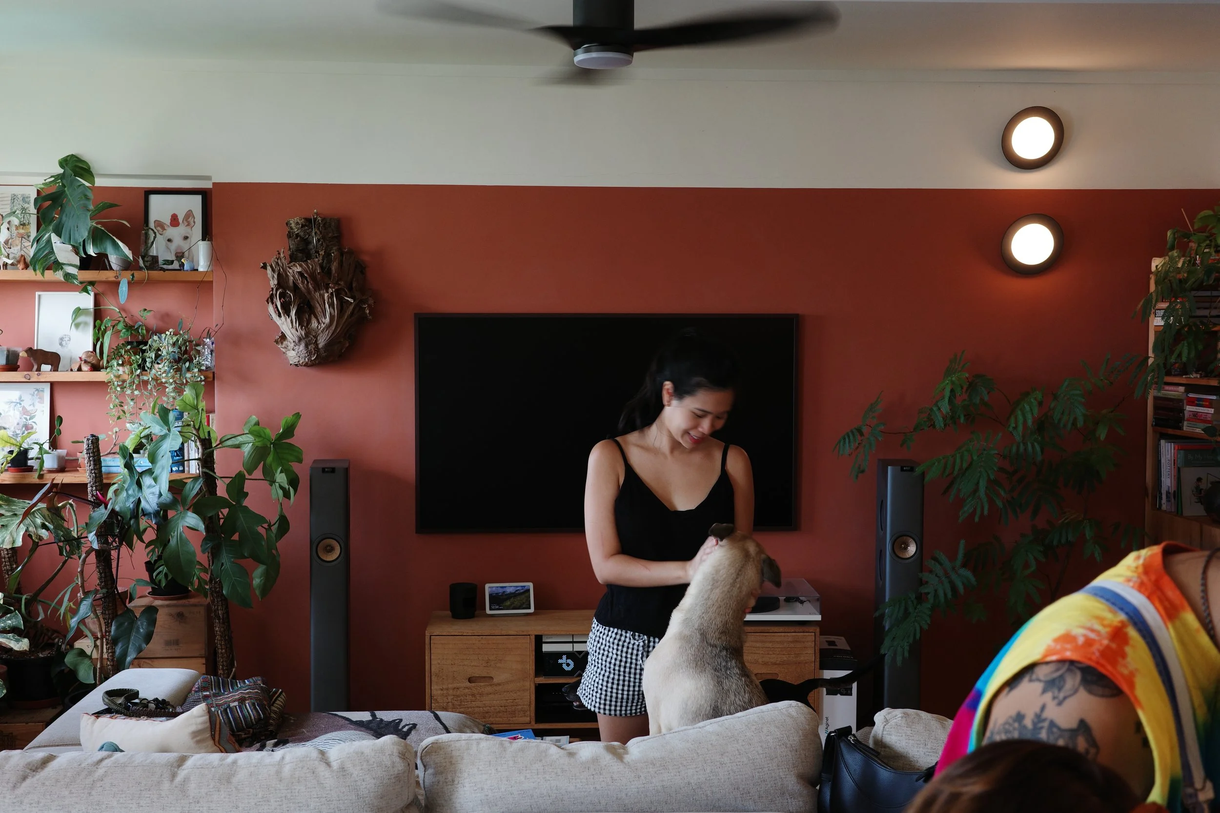 A woman in a black tank top and checkered shorts is indoors, smiling as a dog stands on its hind legs with its paws on her. The background features a red accent wall with a large black TV, plants, shelves with framed pictures and decorative items, an