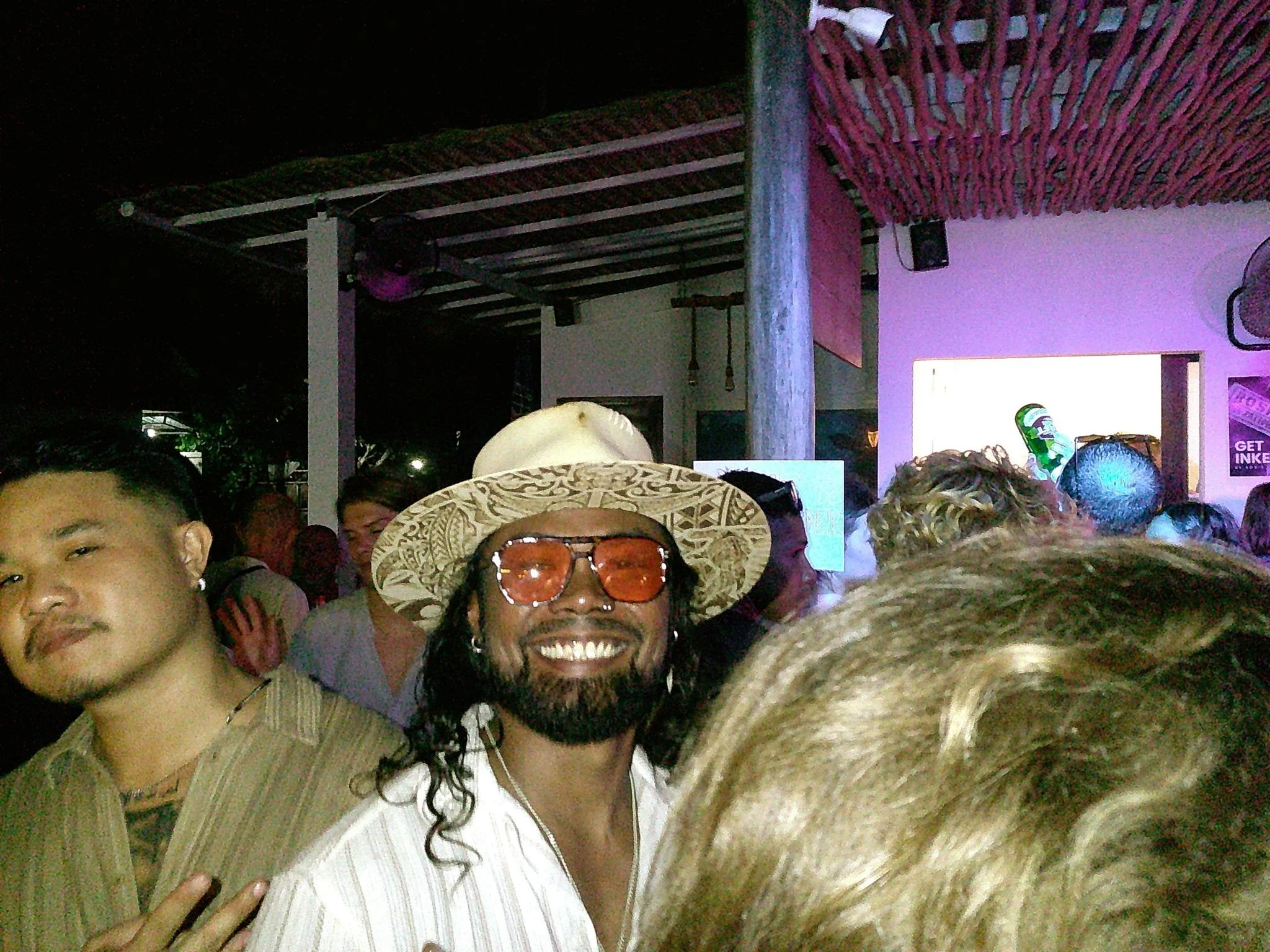 Crowd of people at a party or music event, with a man smiling in the foreground wearing a straw hat and orange sunglasses, and others visible in the background in a dimly lit venue.
