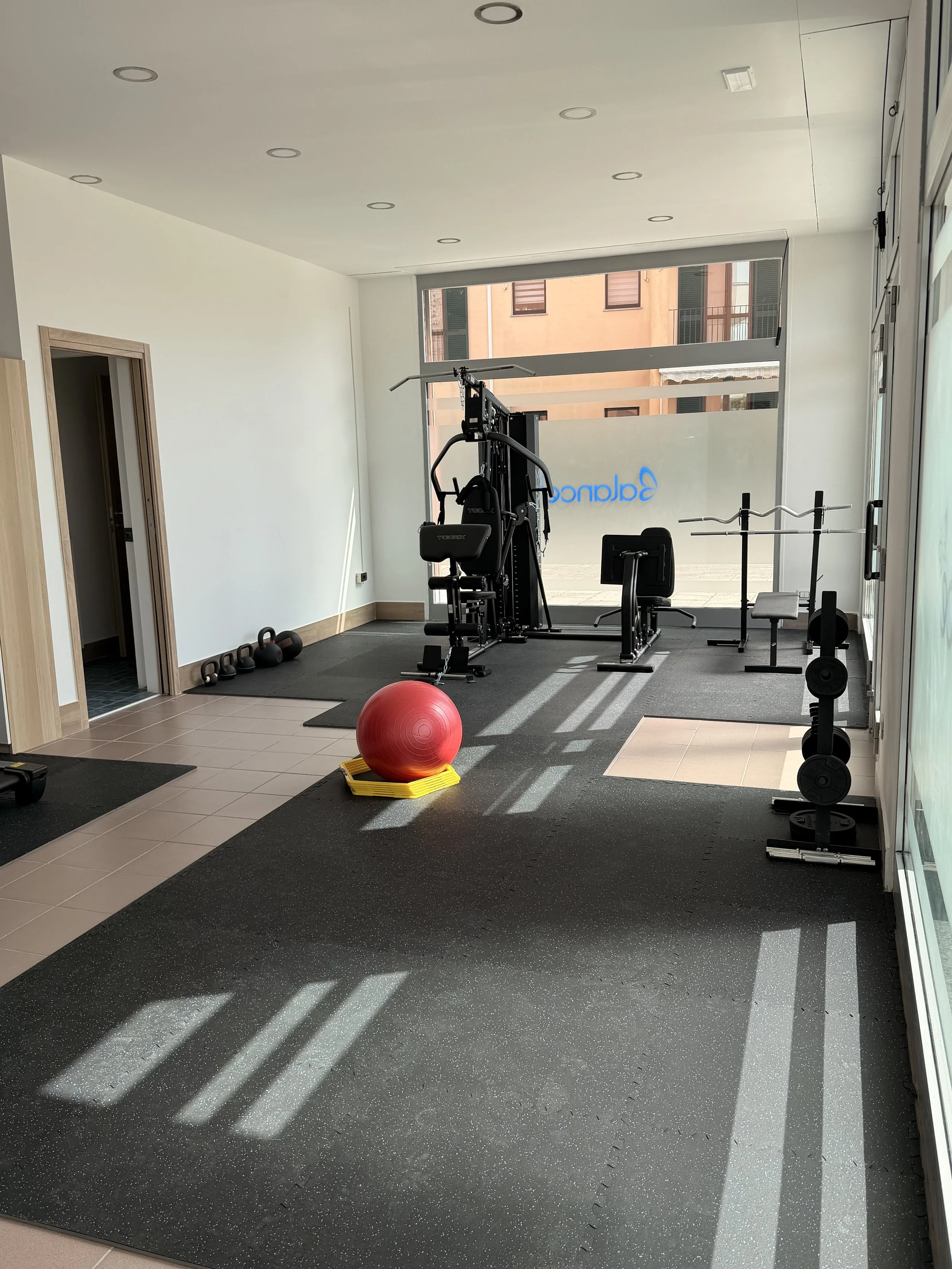 Sala allenamento con attrezzature sportive, panca, squat rack, palla medica rossa, kettlebell, e pesi, con grandi finestre e pavimento in gomma.