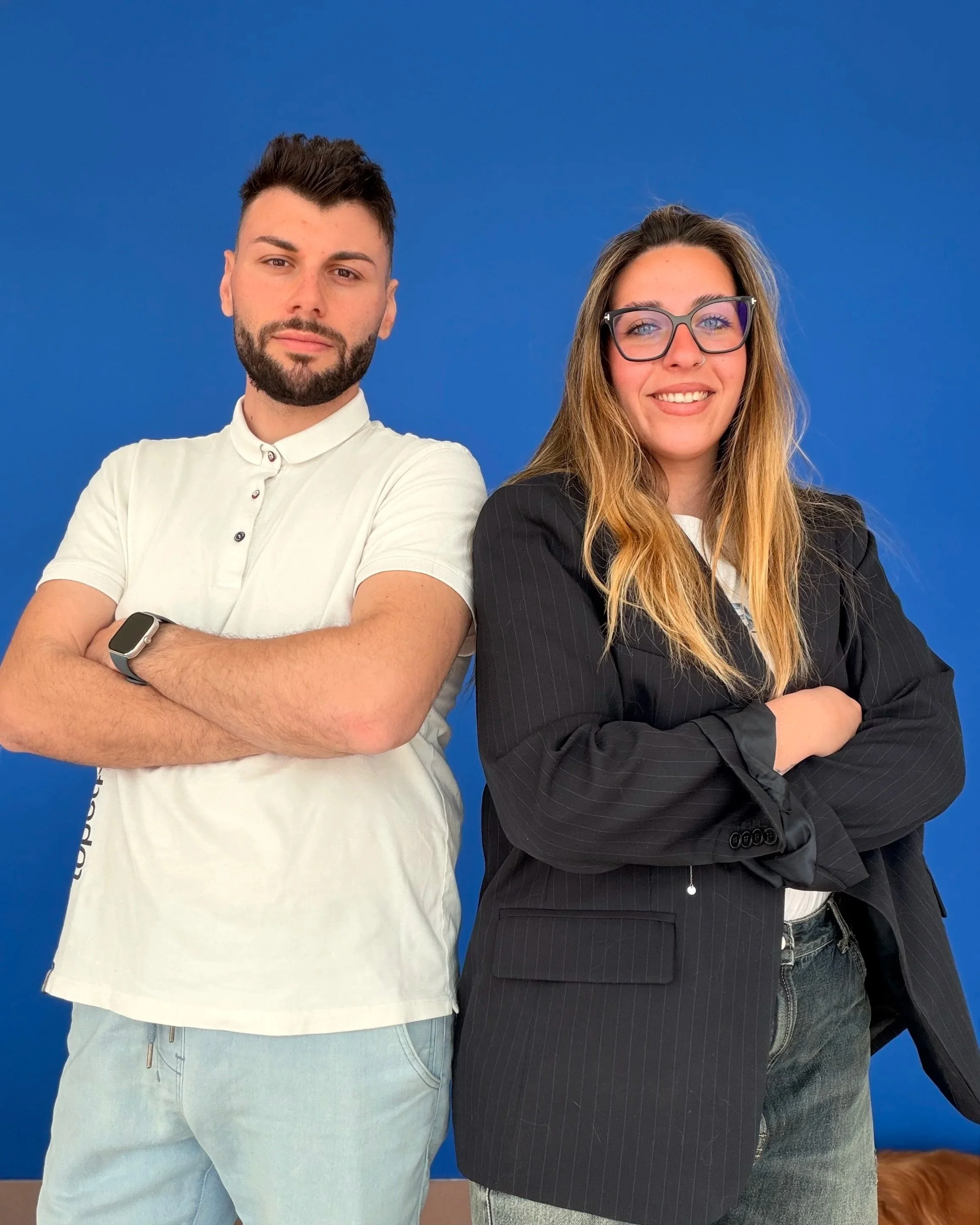 Due giovani adulti, un uomo e una donna, in piedi con le braccia incrociate davanti a uno sfondo blu. L'uomo porta una maglietta bianca e un orologio al polso, ha capelli scuri corti e barba. La donna indossa un giacca nera, occhiali e ha i capelli lunghi di colore castano chiaro. Entrambi sorridono leggermente.