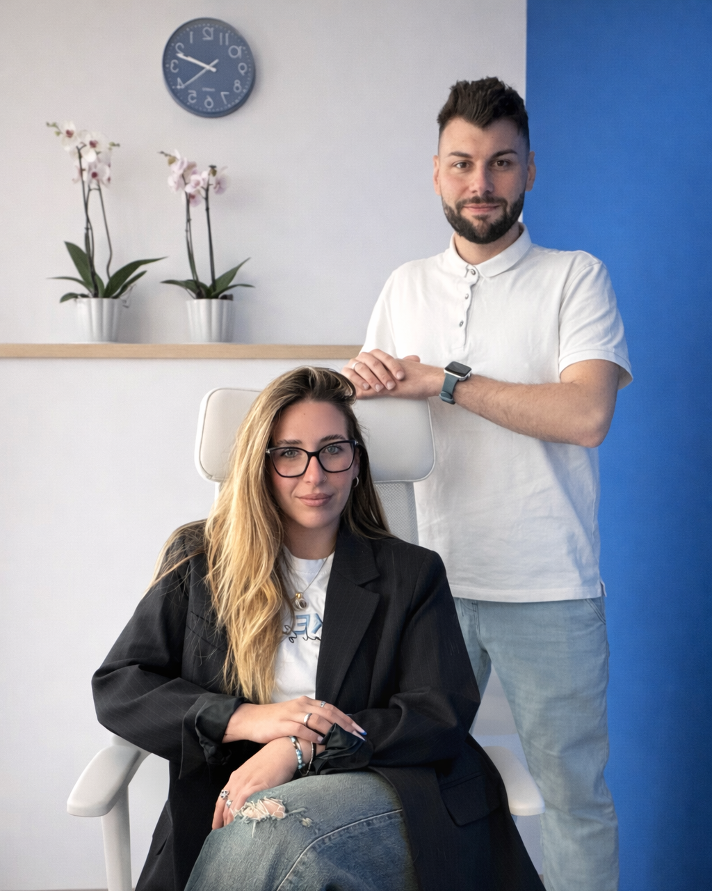 Un uomo e una donna in un ambiente di ufficio. L'uomo in piedi con una maglietta bianca, barba e capelli scuri, si appoggia sulla sedia della donna. La donna seduta, indossa occhiali e una giacca nera, con capelli lunghi e ricci, e jeans strappati.