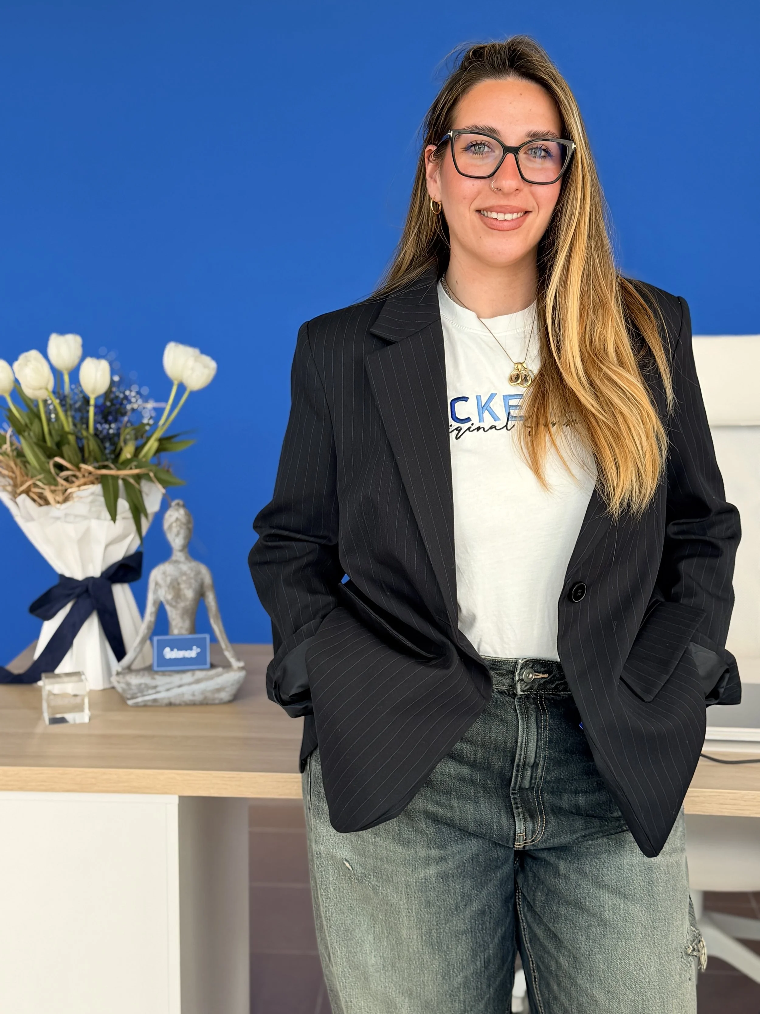 Una donna sorridente con capelli lunghi, occhiali e orecchini d'oro, indossa una felpa bianca e una giacca nera a righe, posiziona le mani nei jeans, in un ufficio con parete blu, fiore di tulipani bianchi e decorazioni sul tavolo alle spalle.