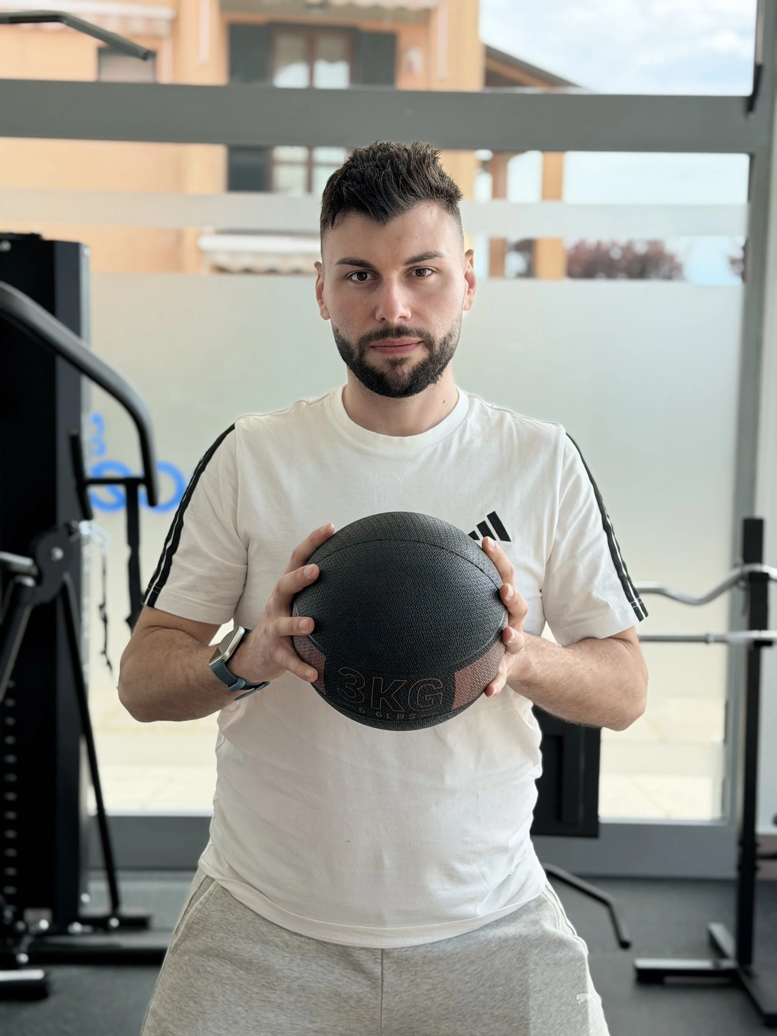Un uomo con barba e capelli corti tiene una palla da esercizio nera in una palestra, con attrezzature sportive visibili sullo sfondo.