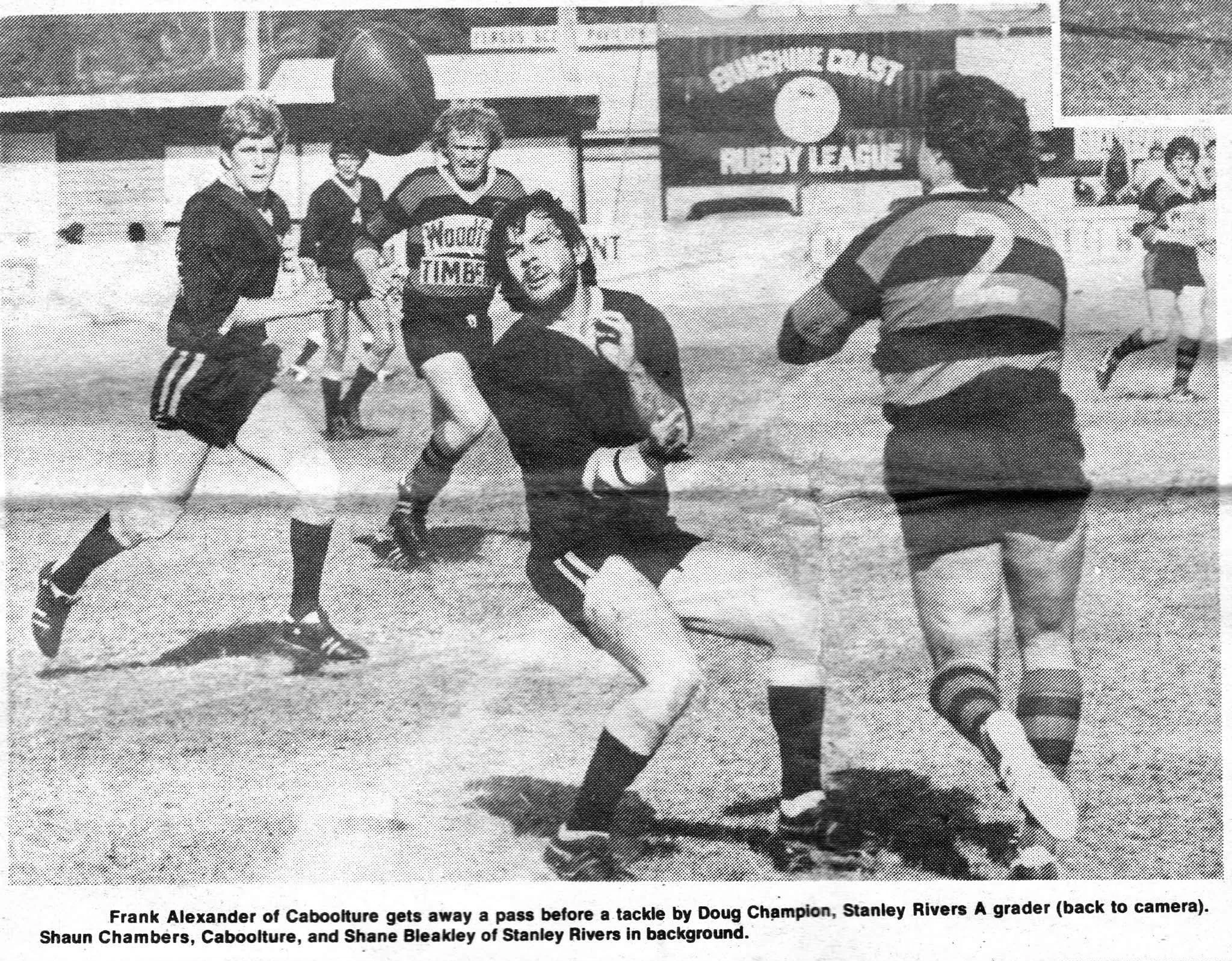 Black and white photo of a rugby game with players from Caboolture and Stanley Rivers teams. A player from Caboolture is running with the rugby ball, attempting to evade a tackle from Doug Champion of Stanley Rivers, who is seen from behind. Other pl