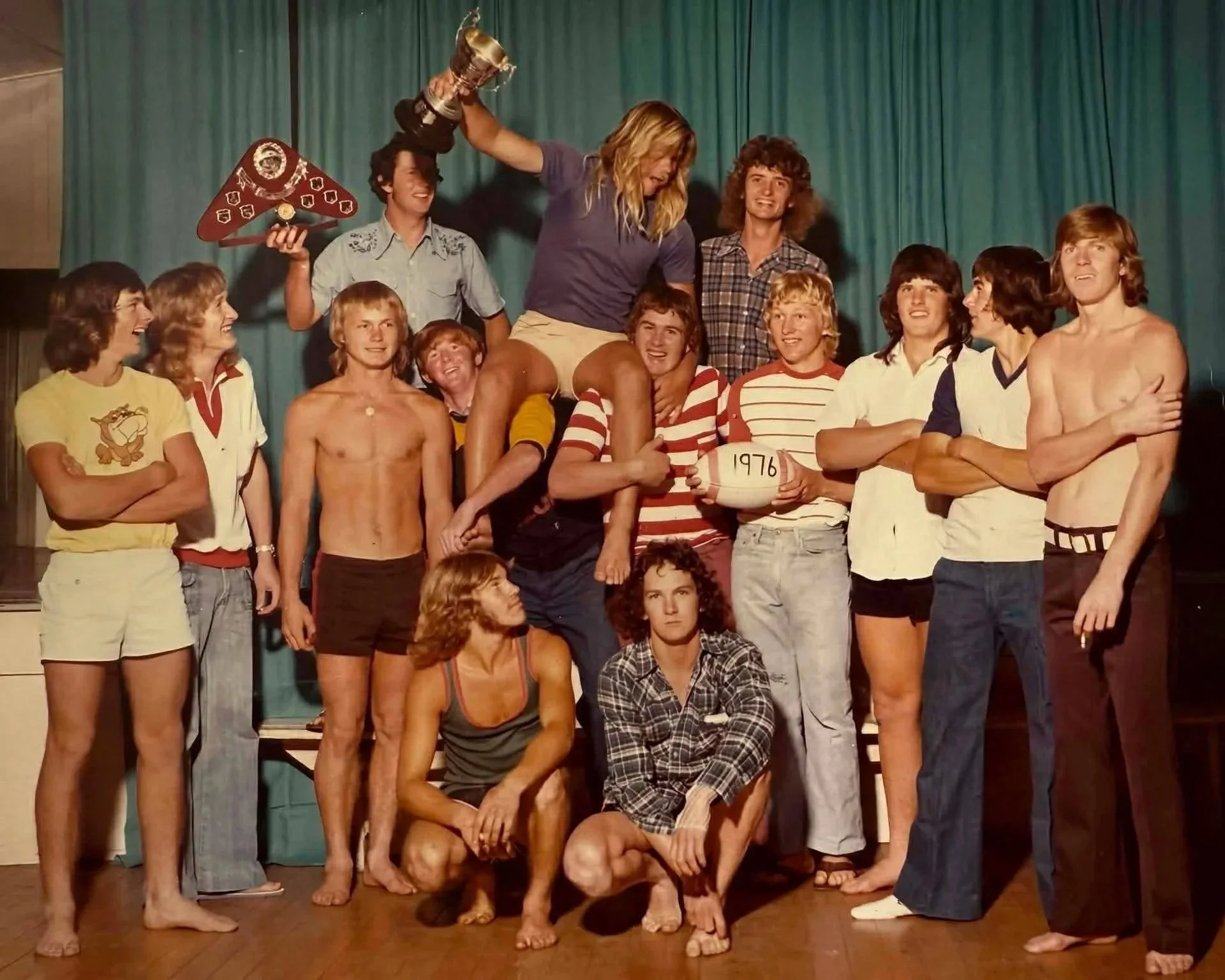 A group of young people on stage celebrating a victory, with one standing on others holding a trophy and a person holding a volleyball marked 1976.