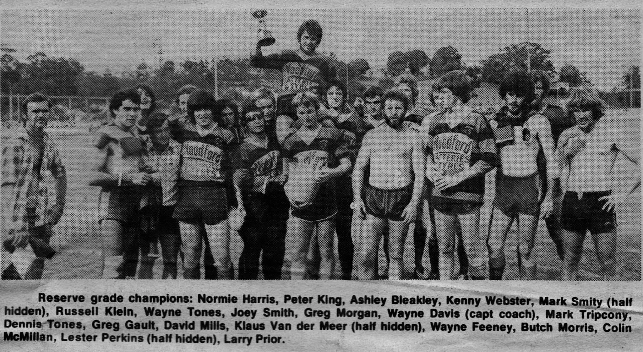A black and white photo of a group of men in sports uniforms, standing outdoors on a field. The caption mentions they are reserve grade champions, and lists the names of some players and the coach.