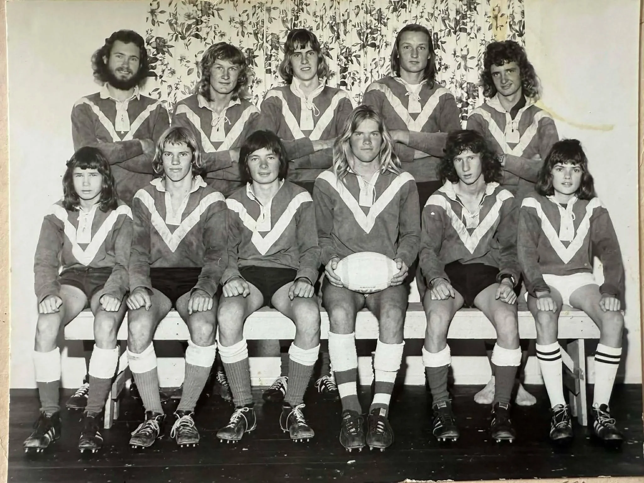 Black and white photo of a rugby team with 11 young women and 2 young men, all in matching uniforms, seated and standing together with a woman holding a rugby ball in the front row, posed in front of curtains.