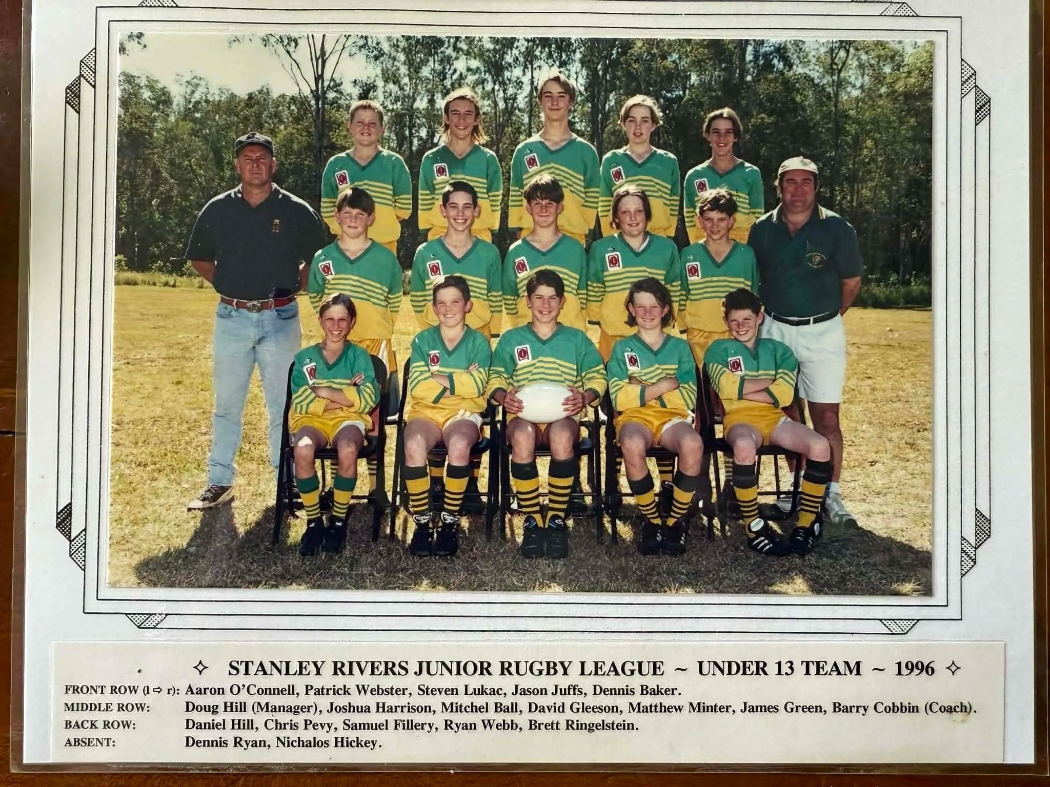 Photograph of 12 young rugby players and 2 coaches from the 1996 Stanley Rivers Junior Rugby League Under 13 Team, posing outdoors on a grassy field with trees in the background, arranged in three rows with the front row seated, the middle row kneeli