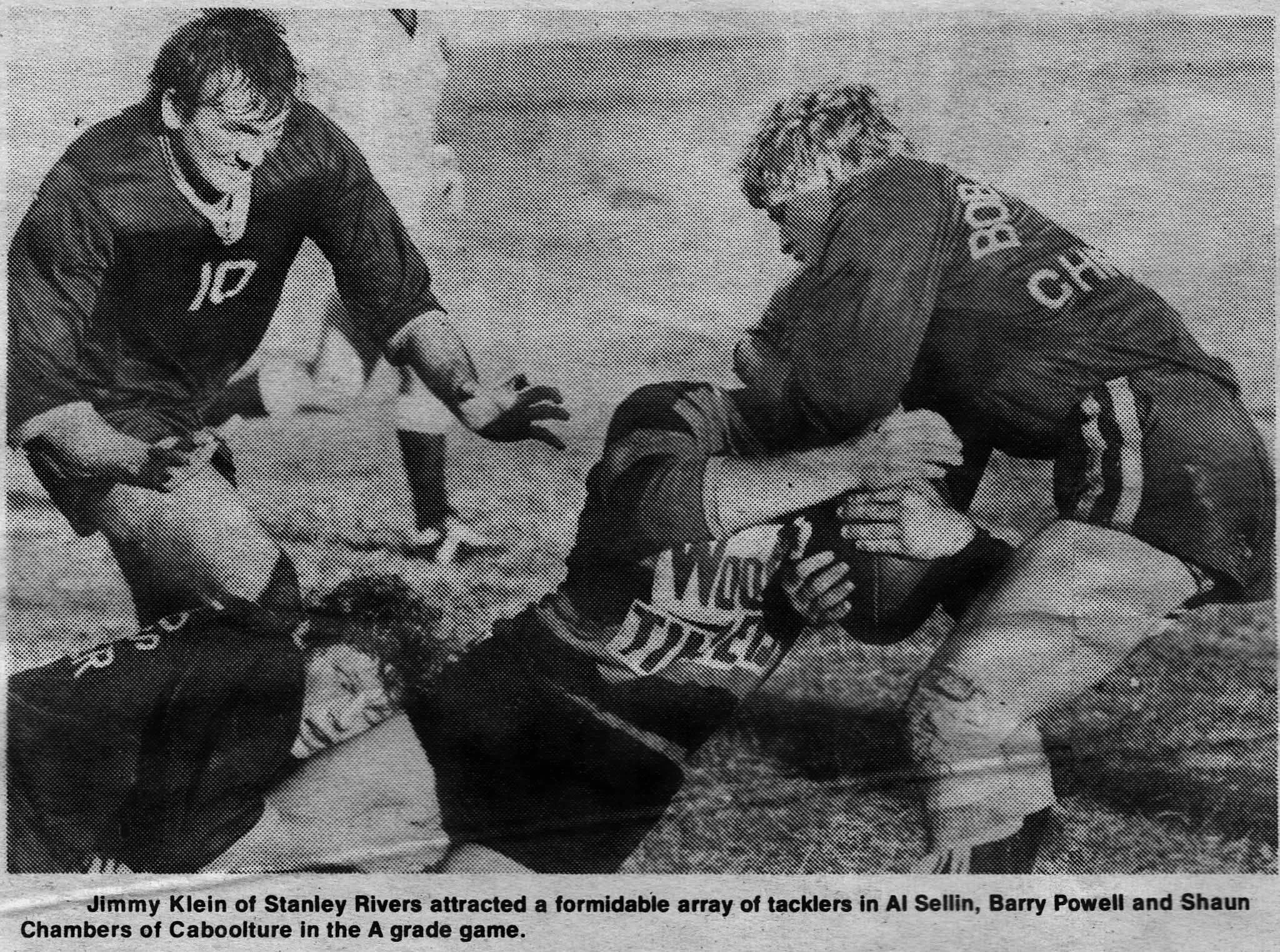 A black and white photo of a football game showing players in action. One player is on the ground while a teammate, Barry Powell, is tackling or helping to tackle an opponent and is being assisted by another teammate, Shaun Chambers. The players are 