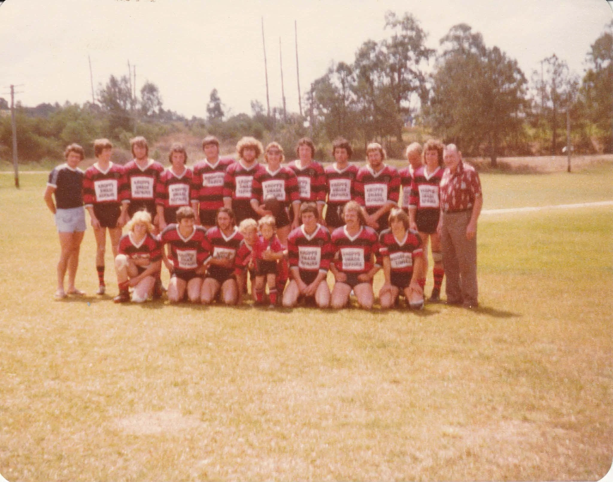 A vintage outdoor photo of a rugby team with players in red and black striped jerseys, kneeling and standing on a grass field, with two women and one man standing at each end.