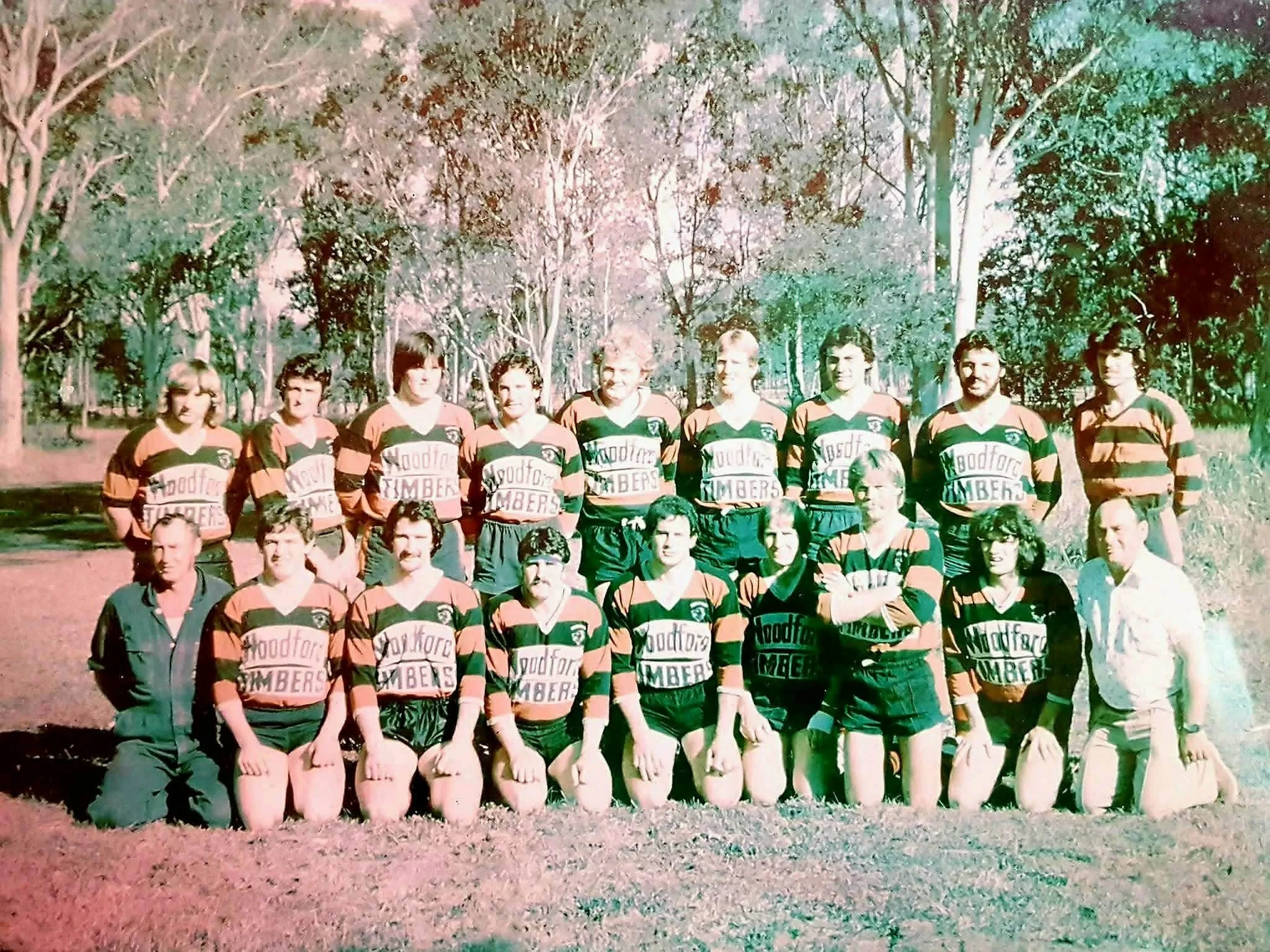 A group photo of a rugby team wearing matching green and red striped jerseys with the sponsor name 'Woodford Timber' printed on the front. They are outdoors in a woodland area with trees in the background. The team is posed in two rows, with some members kneeling in the front and others standing in the back.