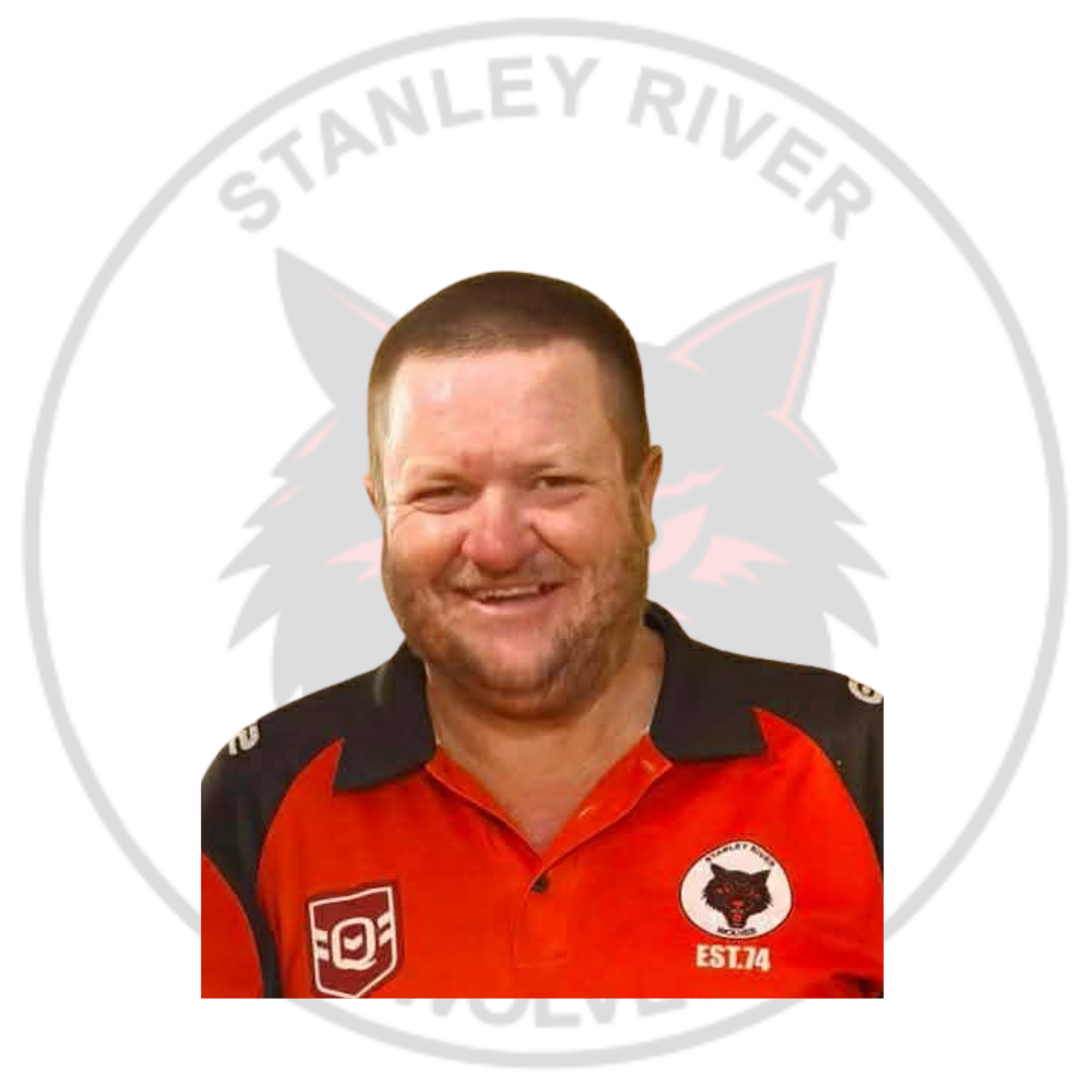 A man smiling outdoors in front of a circular logo that features a black and red wolf's face with the text 'Stanley River' around the top. The man is wearing a red and black sports jersey with a wolf logo on the chest and a patch on the sleeve.