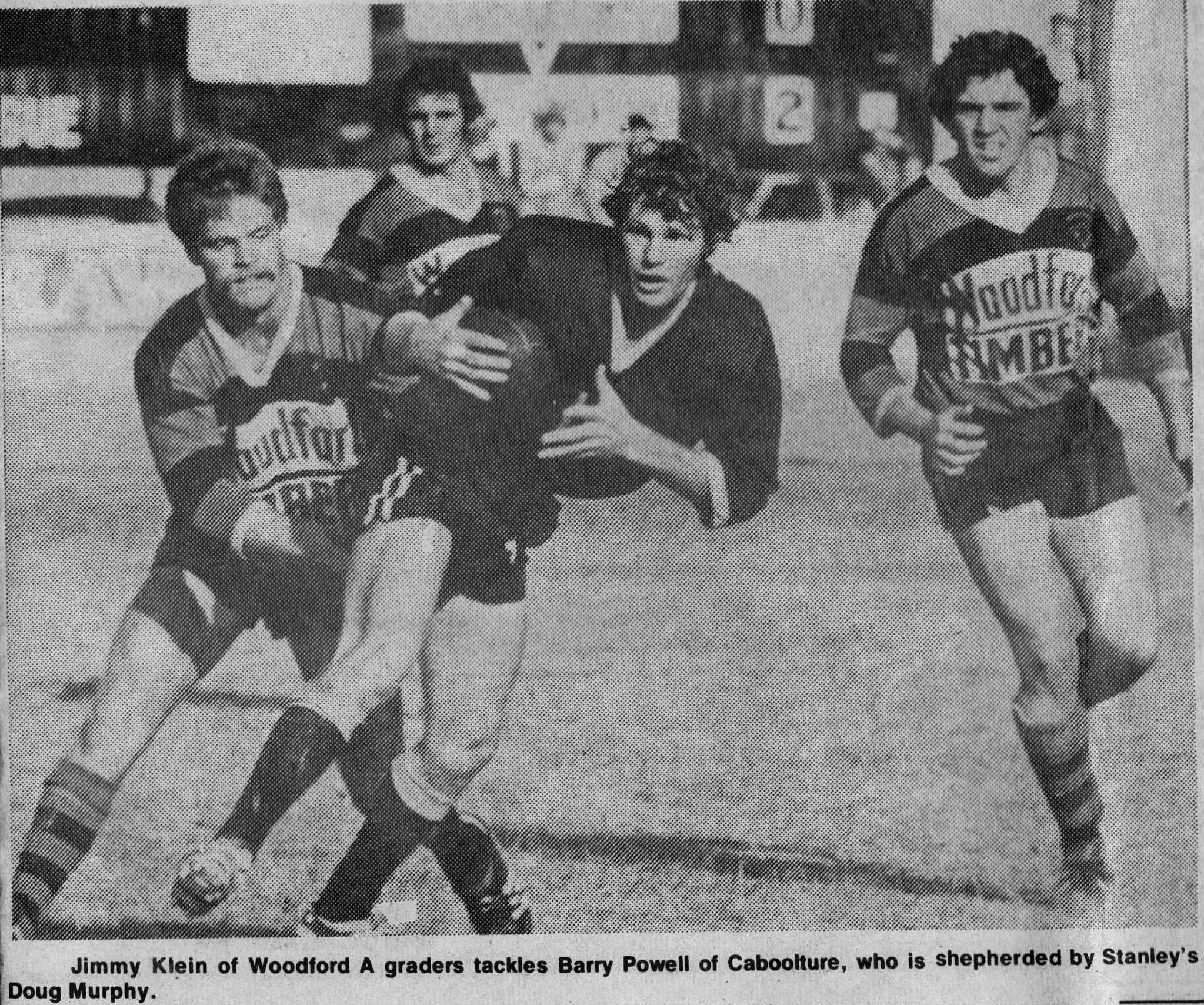 Black and white photo of a football game. A player from Woodford A, Jimmy Klein, is tackling Barry Powell of Caboolture, who is being held by Stanley's Doug Murphy. Klein is wearing a striped jersey, and Powell is carrying the football. Murphy is bes