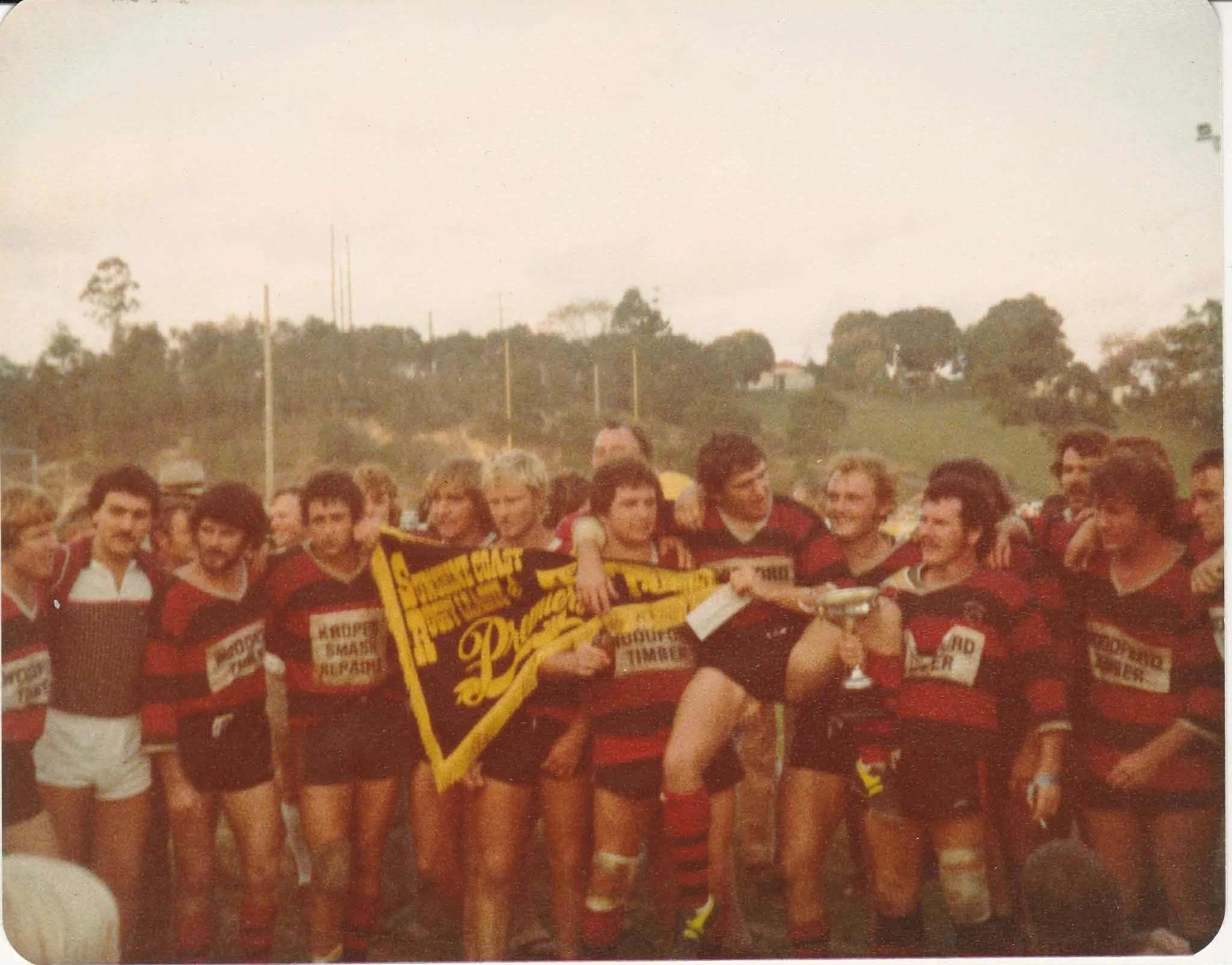 A group of women rugby players in black and red uniforms celebrating together outdoors, some holding a yellow team banner, with trees and hills in the background.