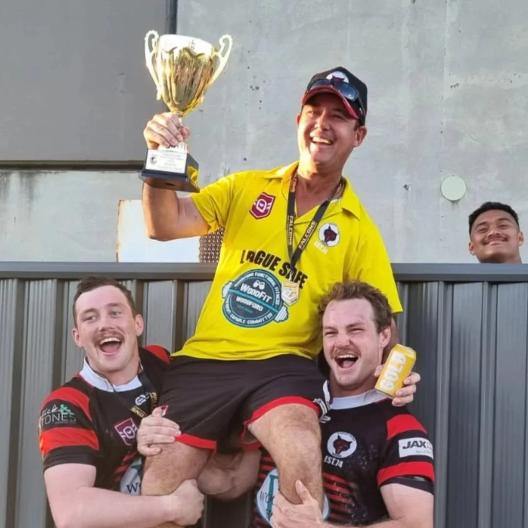 A man in a yellow rugby jersey being lifted by two players in black and red jerseys, holding a trophy and a gold bar, celebrating on a sports field with others in the background.