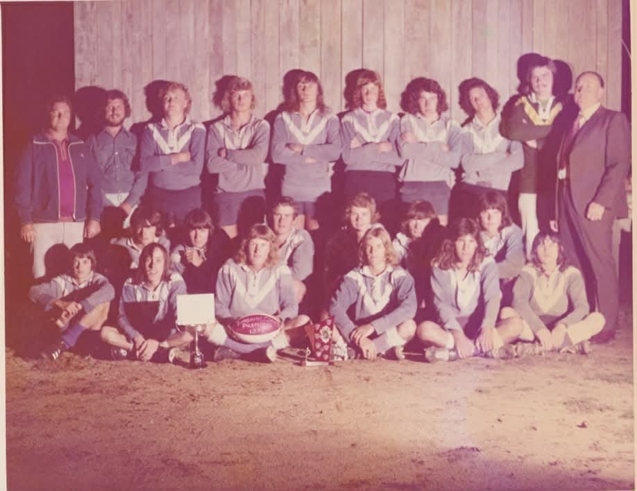 A group of children and three adults posing for a photograph in front of a wooden wall, with some children sitting on the ground and others standing behind them.