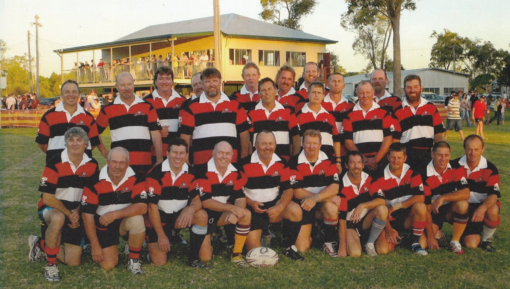 Group of rugby players in red, black, and white striped uniforms posing outdoors with a rugby ball in front, on a grassy field with a building and spectators in the background.