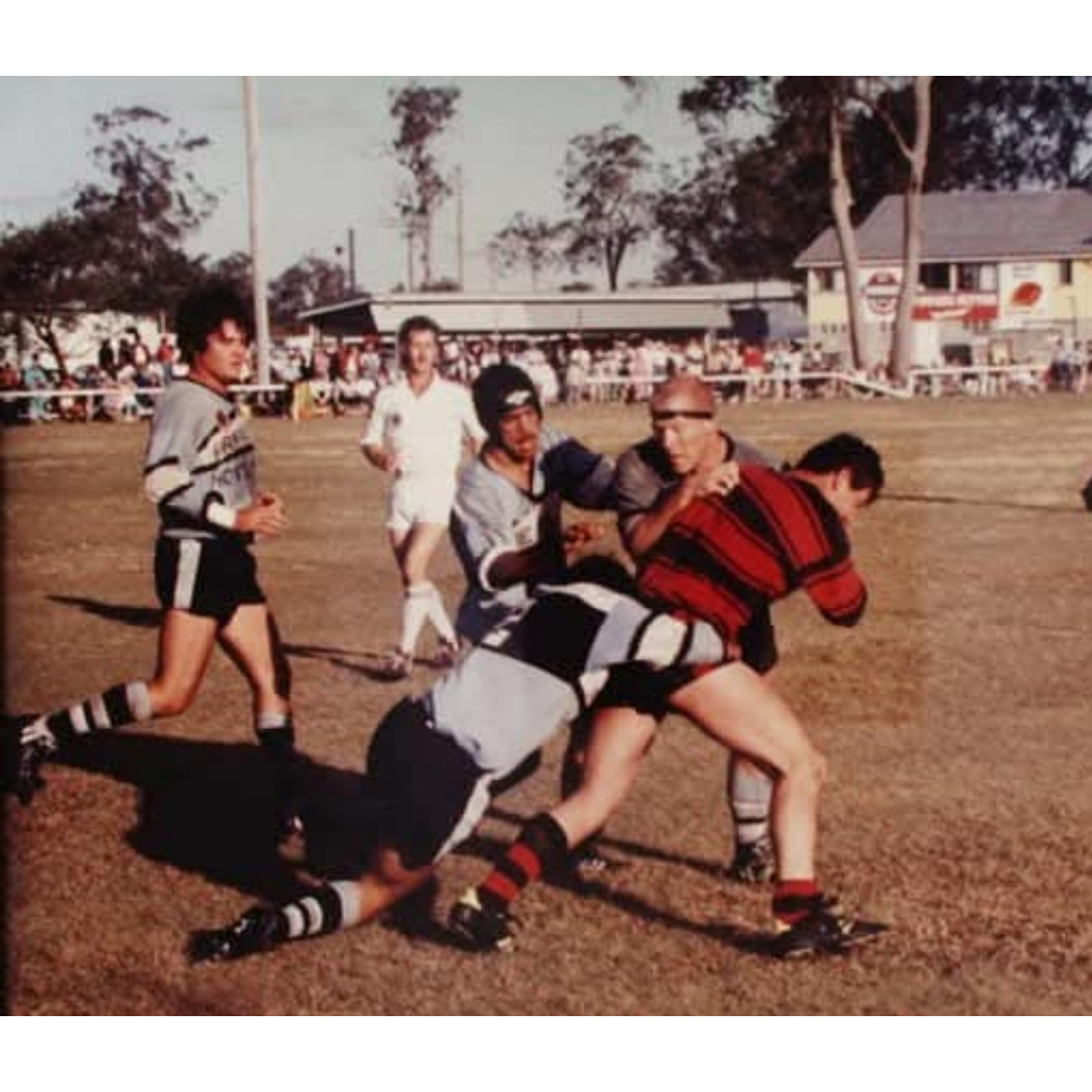 A rugby match with players tackling. One player in a red and black jersey is tackled to the ground by two opponents in black and gray jerseys. Other players and spectators are visible in the background on a dirt field.