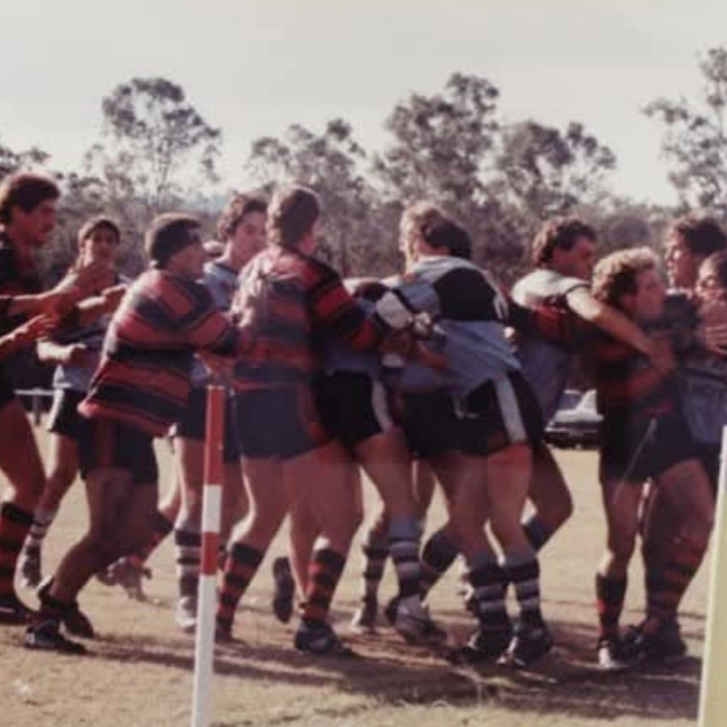 Group of rugby players engaged in a scrum during a game, wearing sports uniforms and knee-high socks, on a grassy field with trees in the background.