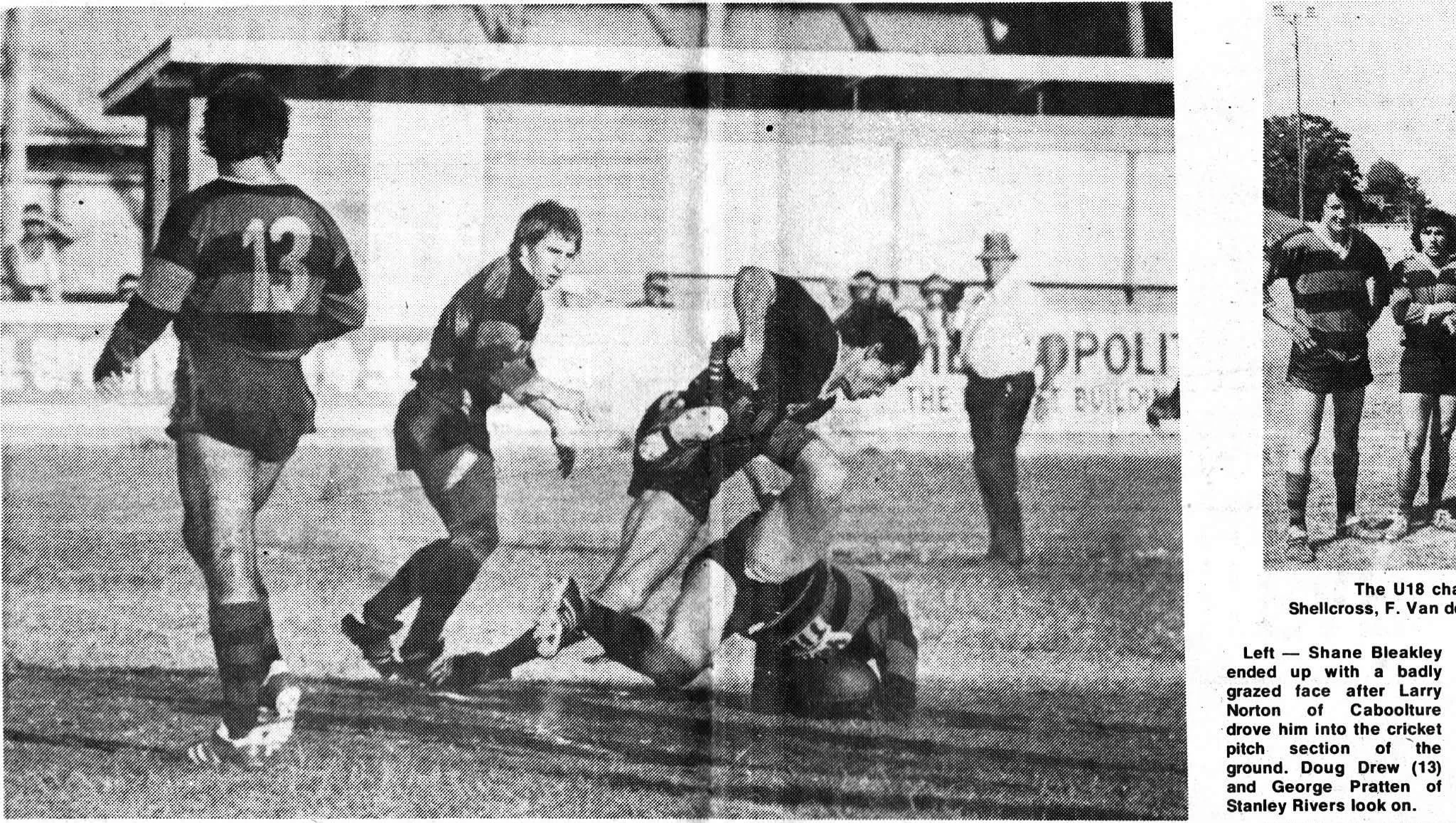 Black and white photo of a rugby match where a player is being tackled by another player on the ground. Two other players and a referee are visible on the field, with spectators in the background.