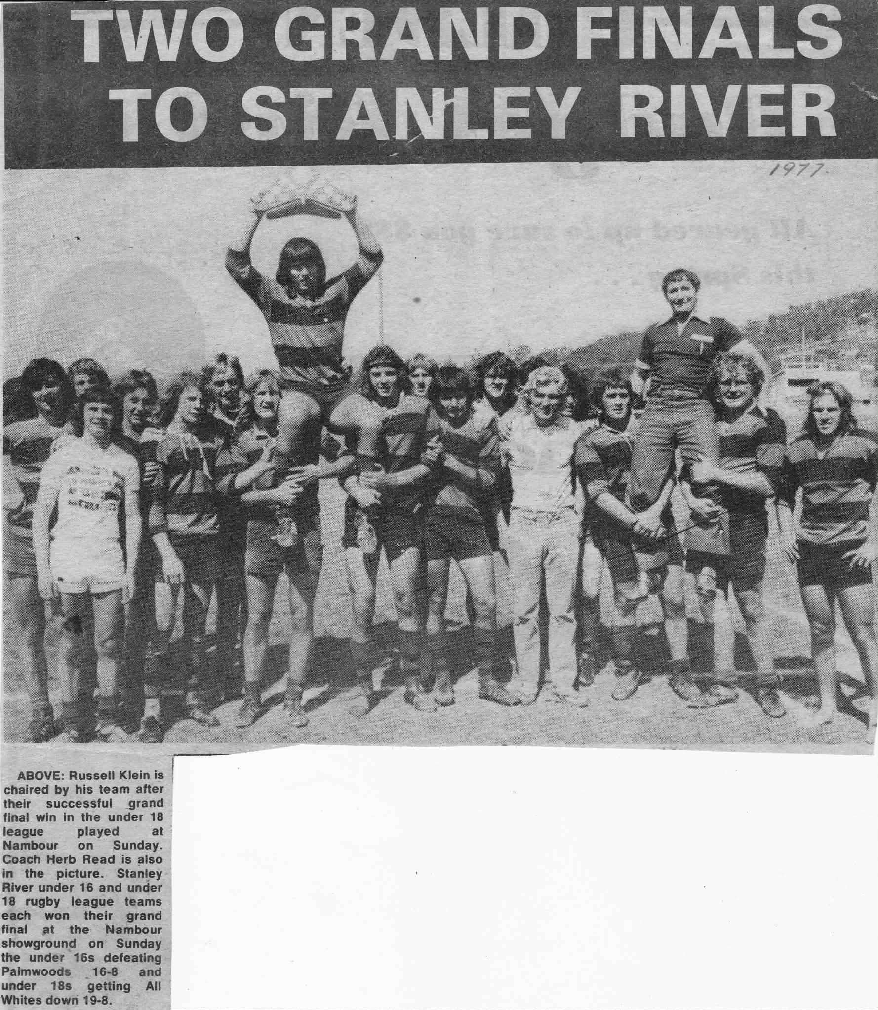 A group of rugby players and coaches, including a woman holding a rugby ball above her head, posing on a field during a sunny day.