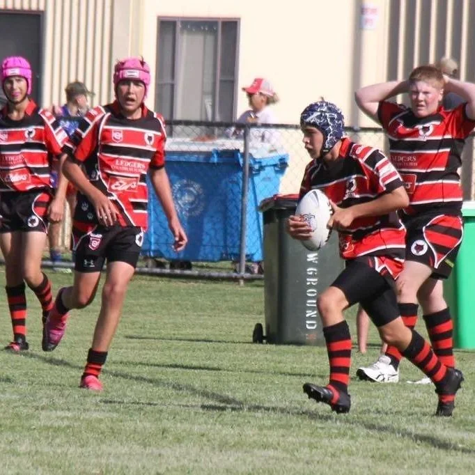 Young girls playing rugby on a field, wearing matching red, black, and white uniforms, some with pink or blue hats and protective headgear, one girl holding a rugby ball and running.