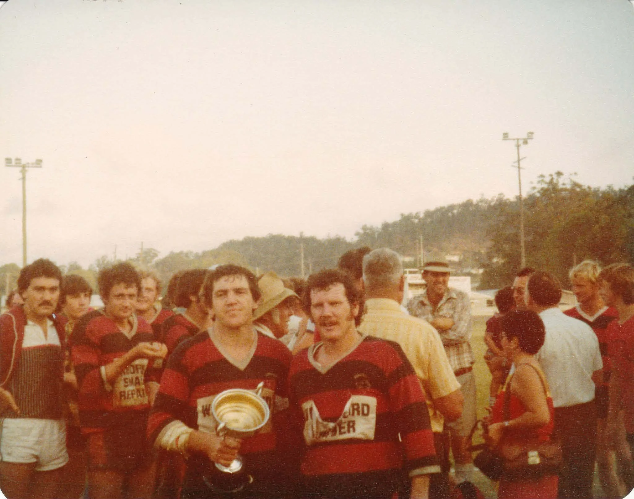 A vintage photograph of a rugby team holding a trophy, with players wearing red and black striped jerseys, gathered outdoors on a field with spectators and trees in the background.