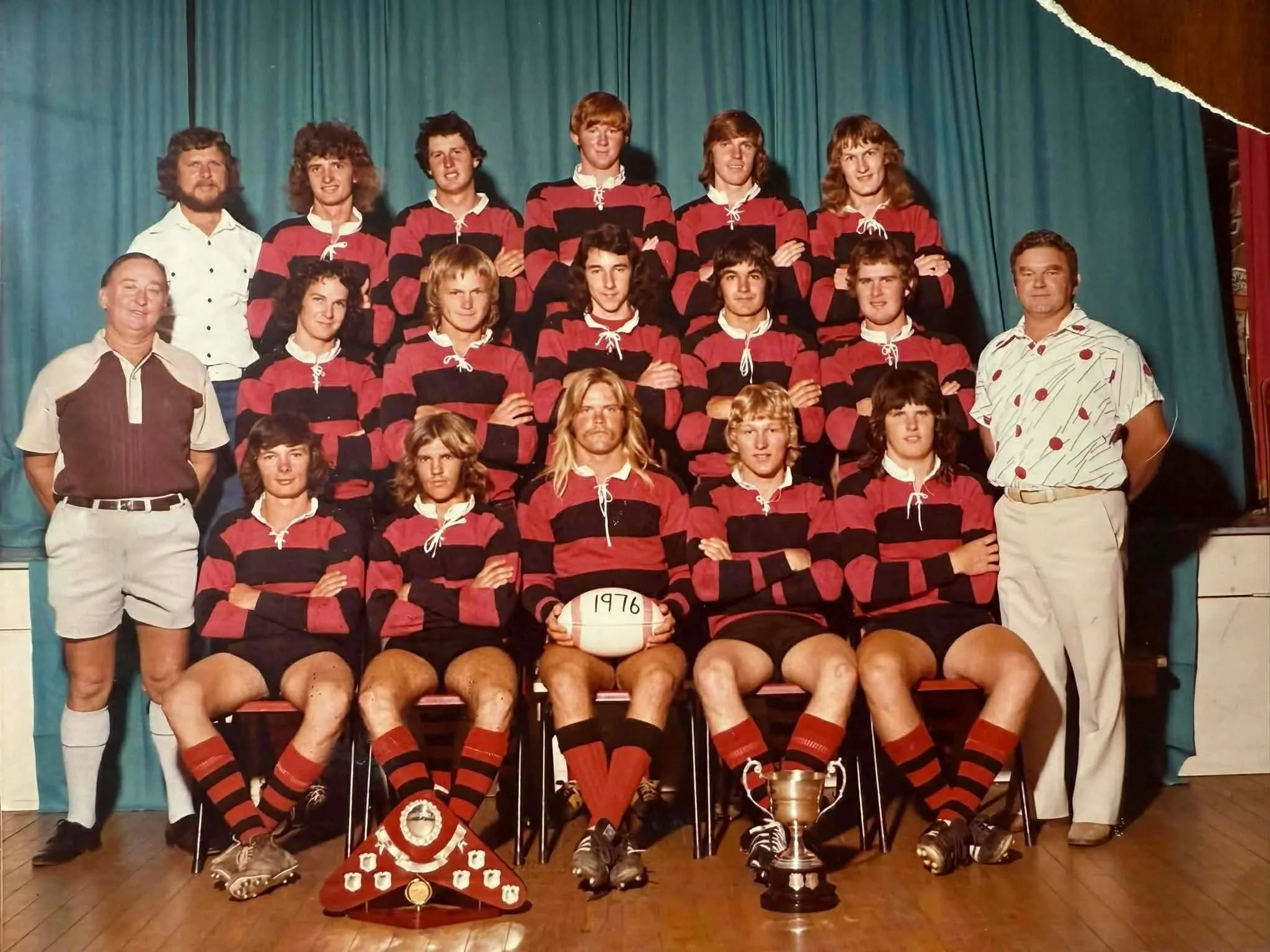 A rugby team posing for a group photo in 1976, with players wearing red and black striped uniforms, two coaching staff in casual attire, and trophies in front