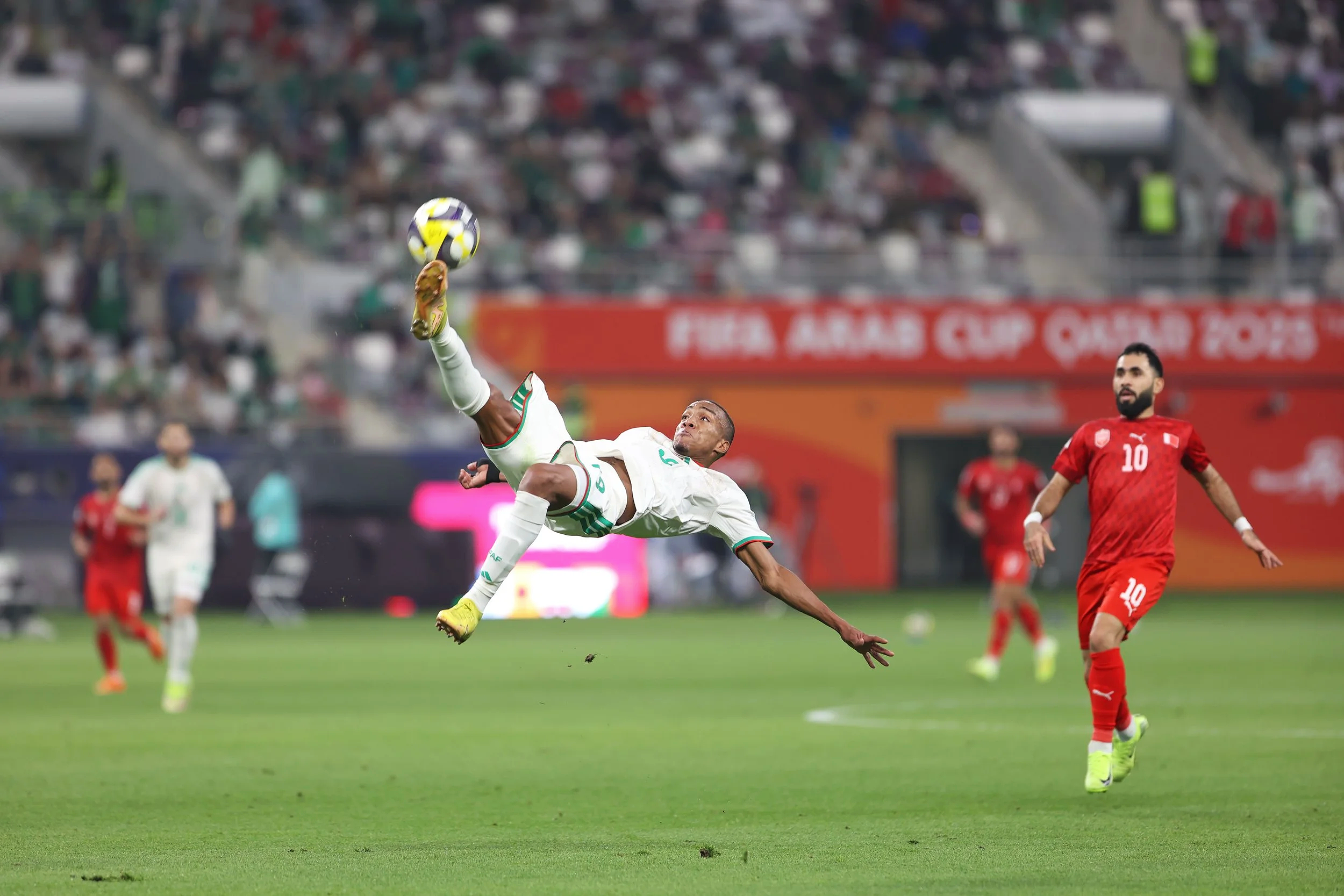 A soccer player in a white jersey is mid-air executing a bicycle kick to hit the ball, while other players and spectators observe on the field during a match.