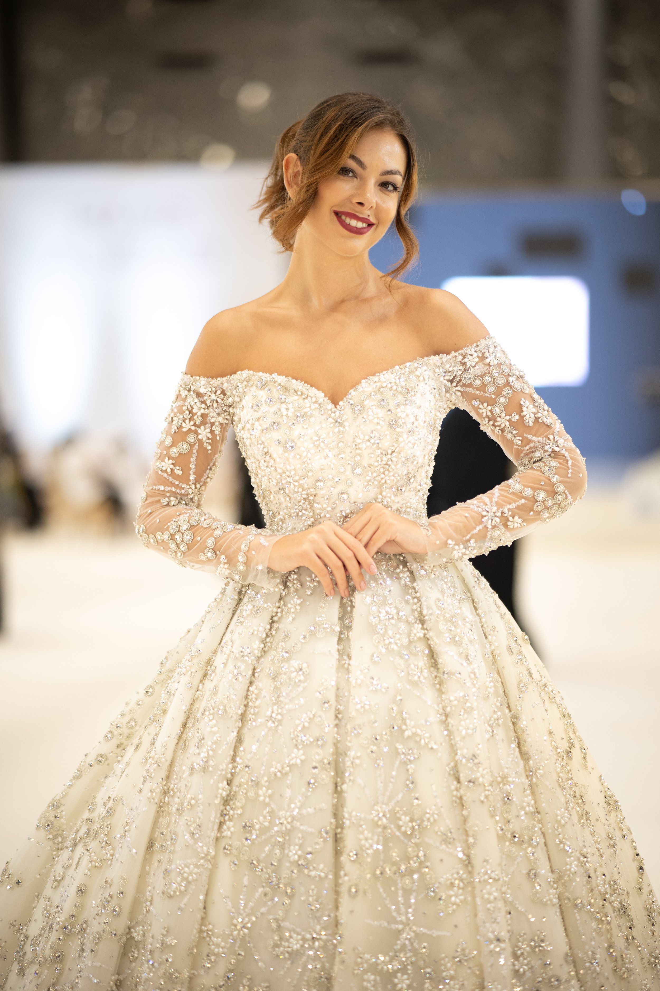 Woman wearing an ornate off-shoulder wedding gown with lace and bead embellishments, smiling with hands clasped, indoors with blurred background.