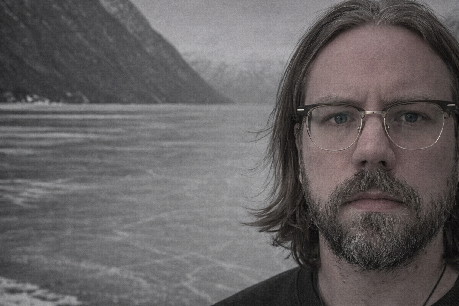 A man with shoulder-length hair, glasses, and a beard stands near a body of water with mountains in the background, in a grayscale or muted color photo.