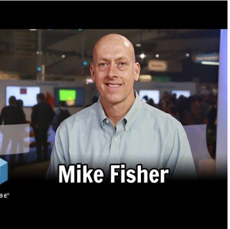 A man with a shaved head and a light blue button-up shirt smiling at the camera, with a busy technology or conference event in the background.