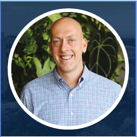 A smiling man with a bald head wearing a blue checkered shirt, standing in front of green foliage.