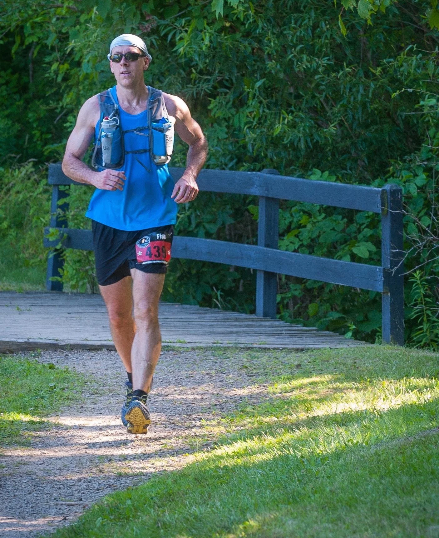 A man running on a trail surrounded by greenery, wearing a blue sleeveless shirt, black shorts, sunglasses, a white headscarf, and a hydration vest.