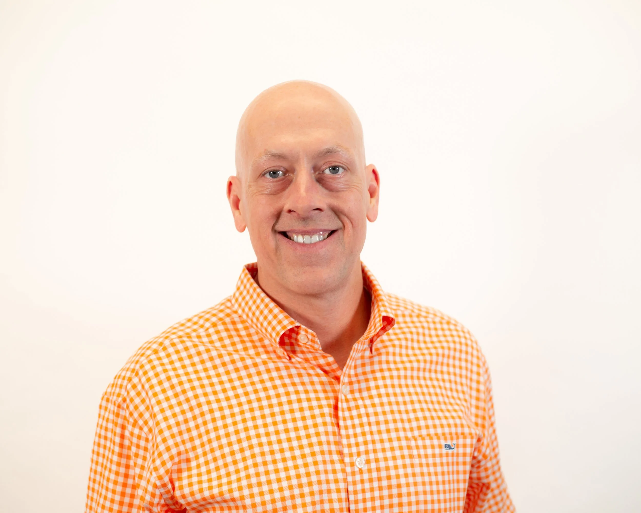 Headshot of a smiling man with a shaved head wearing an orange and white checkered shirt against a white background.