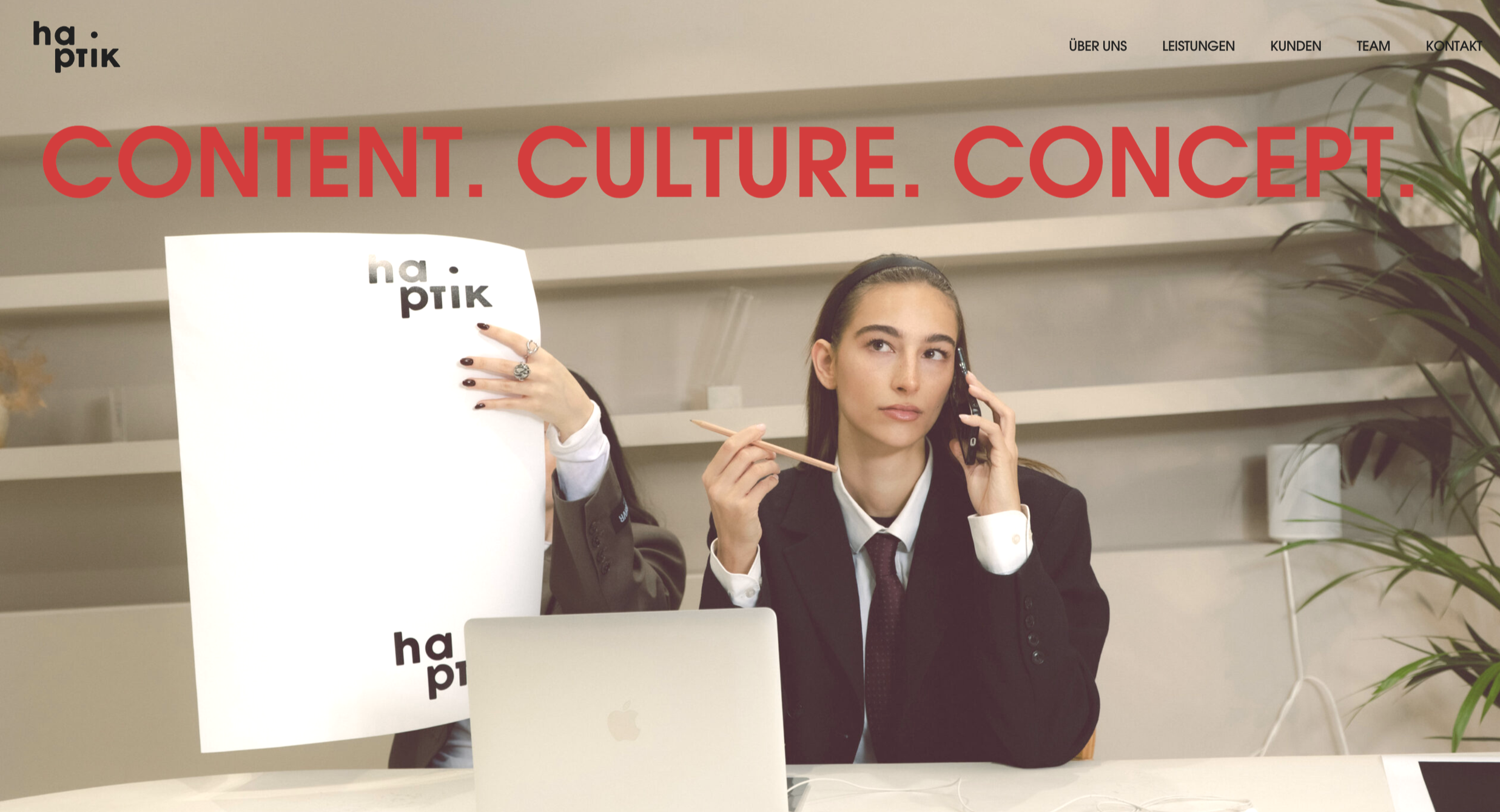 A woman in business attire is seated at a desk, holding a phone to her ear with her right hand and a pencil in her left. Behind her, someone is holding a large poster with the logo ‘ha.pptk’. The background shows a modern office with white walls, she